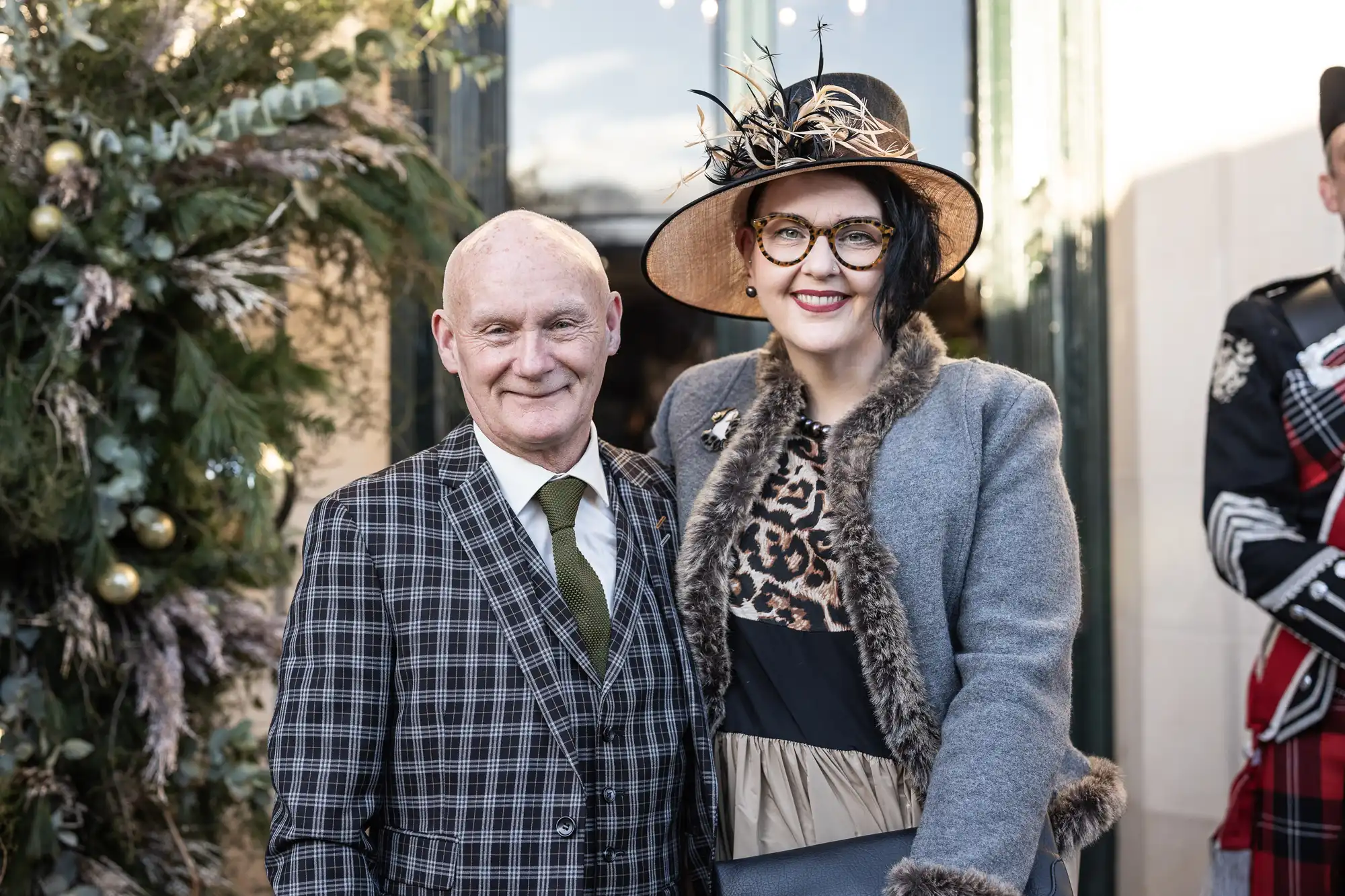 A man and woman dressed in formal attire stand together outdoors. The man wears a checkered suit; the woman wears a hat and a gray coat with a leopard print top. A person in traditional attire stands behind them.