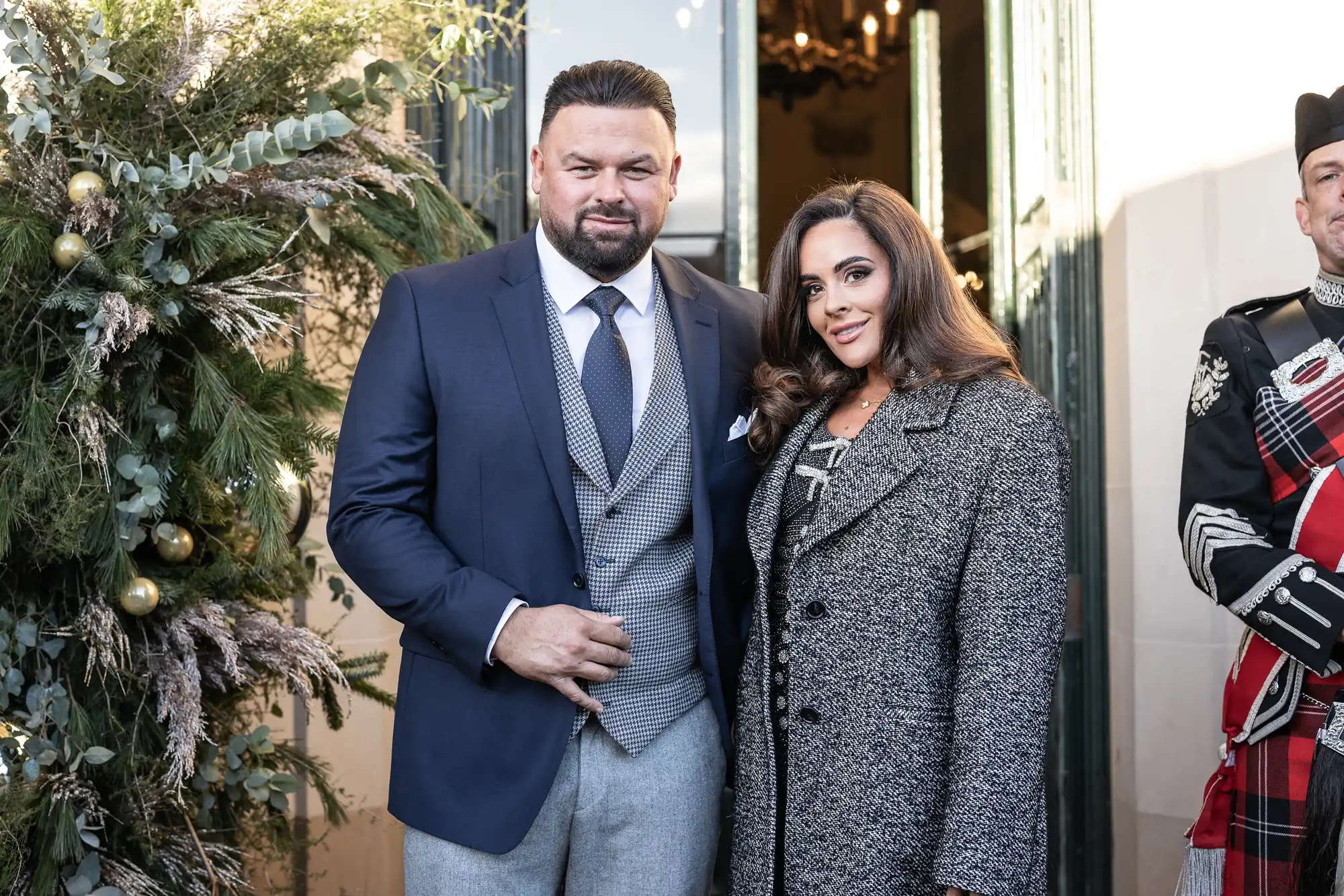 A man in a suit and a woman in a coat stand outdoors, smiling for the camera. A man wearing traditional attire is partially visible on the right. Greenery decorates the background.