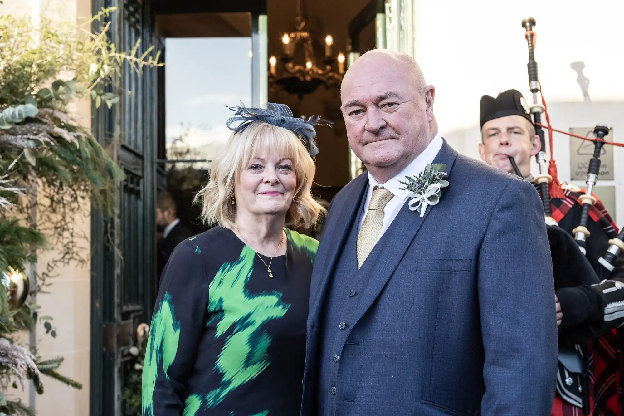 A man and woman, dressed formally, stand together at an event. A bagpiper is in the background. The woman wears a patterned dress and fascinator, and the man wears a suit with a boutonnière.