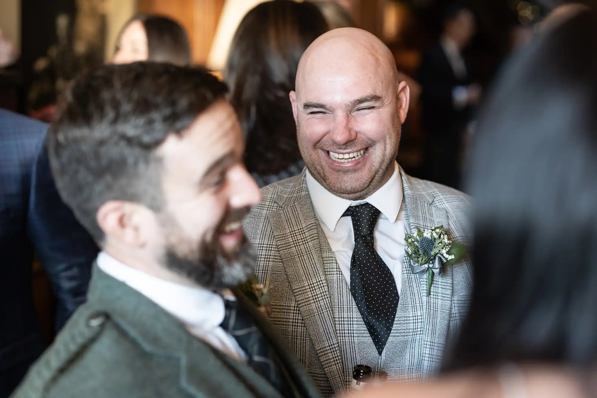 Two men in formal attire are laughing and conversing at an indoor social gathering. One is in a plaid suit with a boutonniere, and the other is in a dark suit with a tie. People are visible in the background.