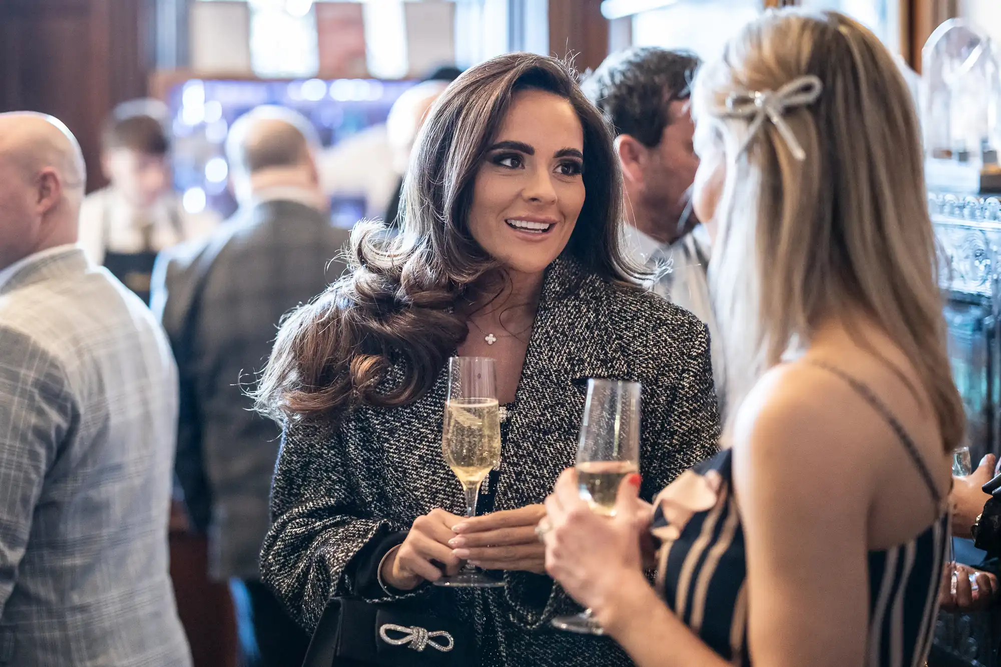 Two women holding glasses of champagne converse in a bustling social setting with other attendees visible in the background.