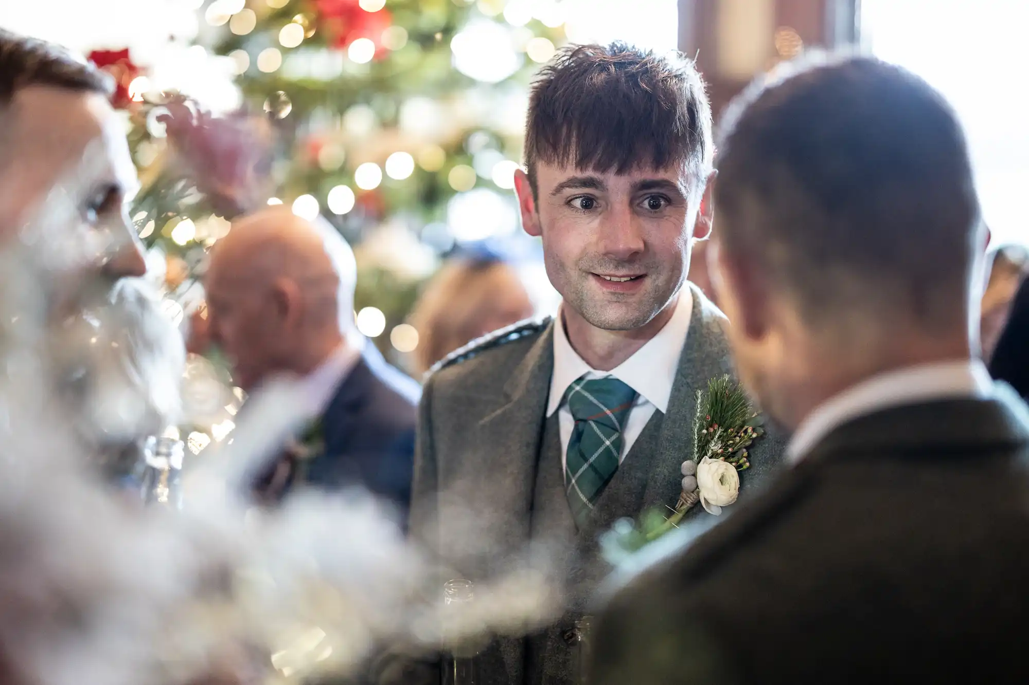 A man in a grey suit and tartan tie stands and talks to another person at a social gathering. A decorated Christmas tree is in the background.