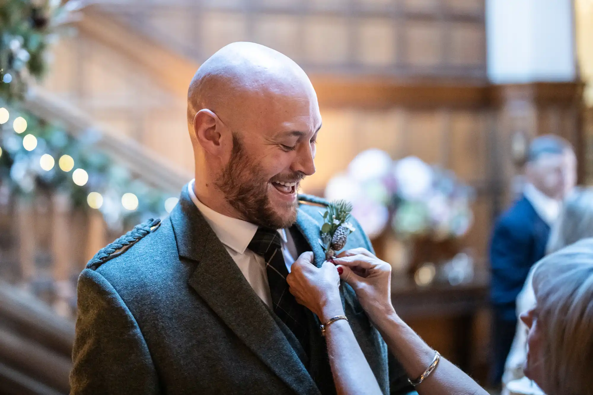 A bald man in a suit smiles as another person adjusts his boutonniere. They are in a warmly lit, wood-paneled room adorned with festive decorations.