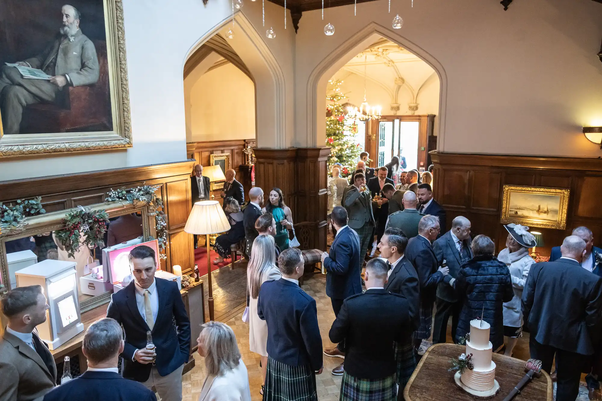 A group of people, dressed in formal attire, gather in a wood-paneled room decorated for Christmas with a tree and lights. A few are engaged in conversations, while others stand or move around.