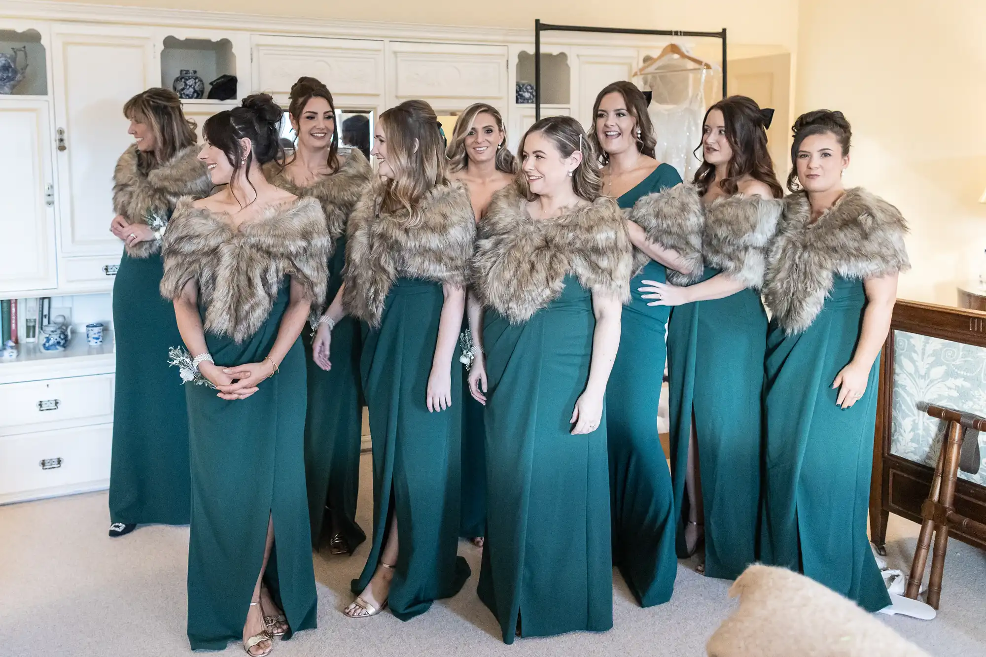 A group of nine women wearing matching green dresses with faux fur stoles stand together in a room, smiling and looking in different directions.