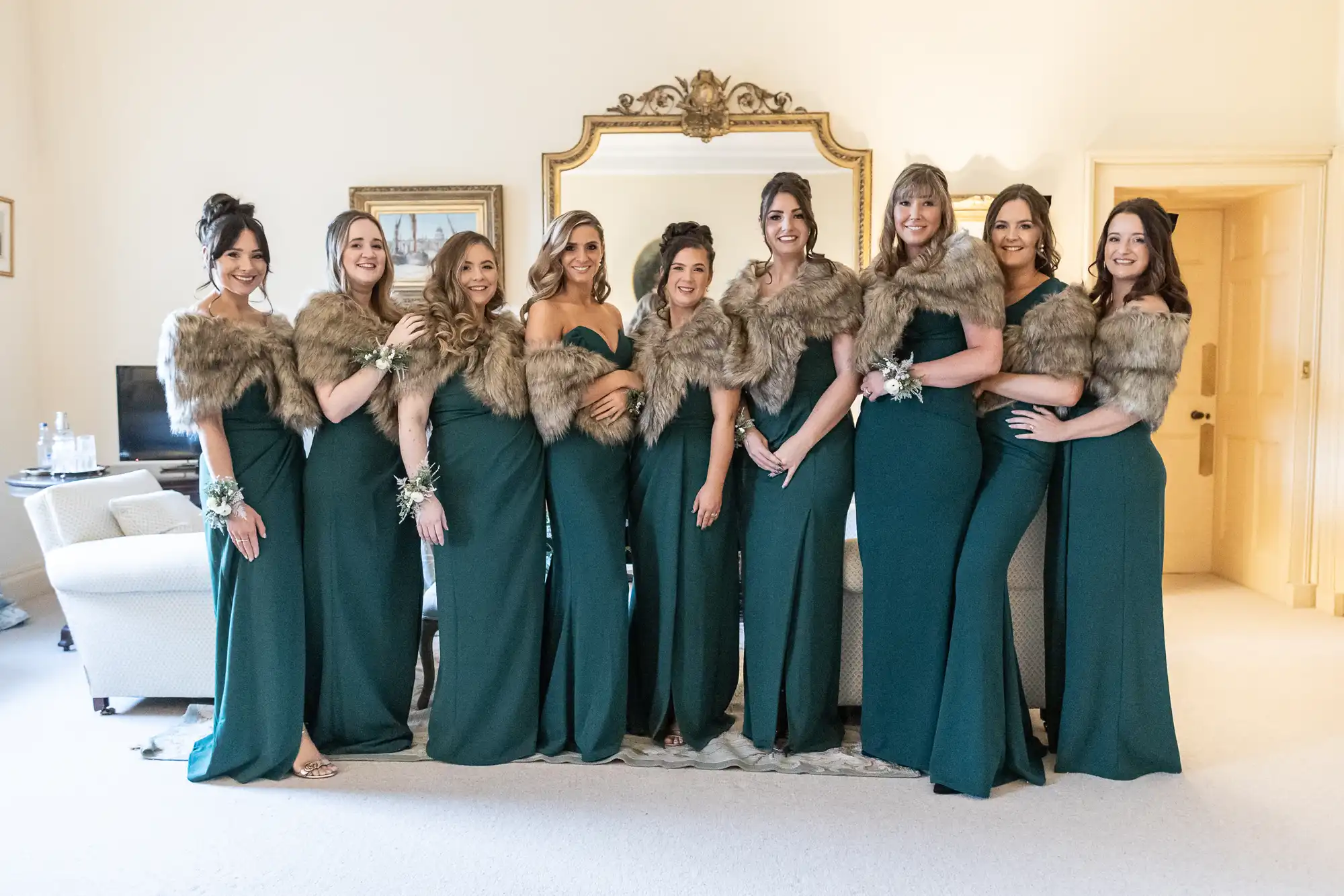 A group of nine women dressed in matching green gowns with fur wraps stand in a line in a room with ornate mirrors and furnishings.