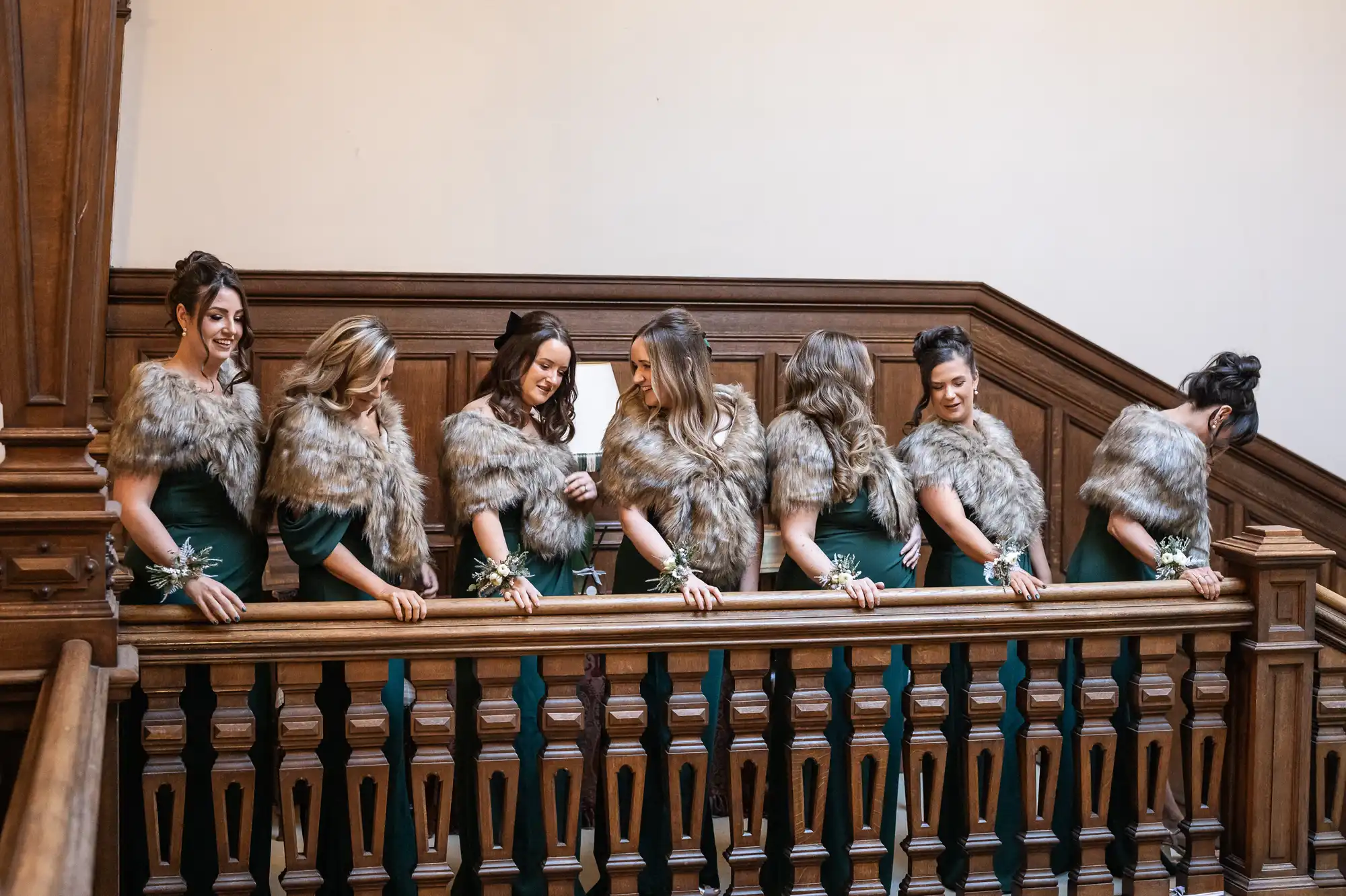 Six women dressed in green gowns and faux fur shawls stand in a line on a wooden staircase, facing slightly towards each other.