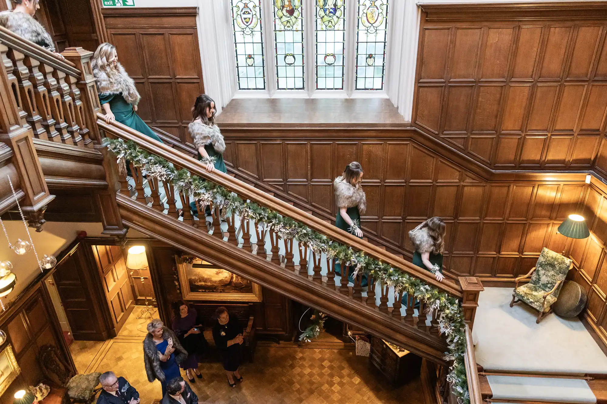 Several people are descending a wooden staircase adorned with greenery, in a room with wooden paneling and a large, stained glass window. A few individuals are seen standing on the ground floor.