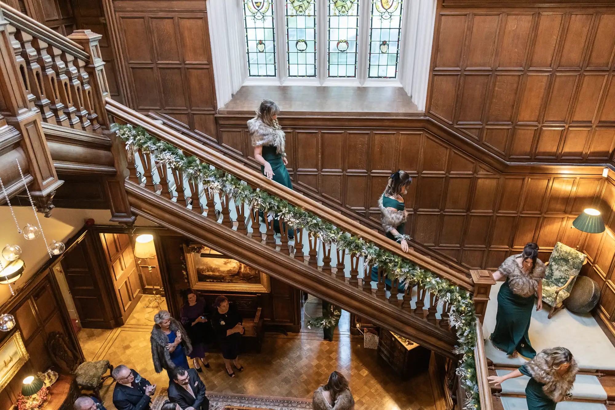 A group of women in green dresses descends a wooden staircase adorned with garland, while others gather on the floor below in a wood-paneled room with stained glass windows.