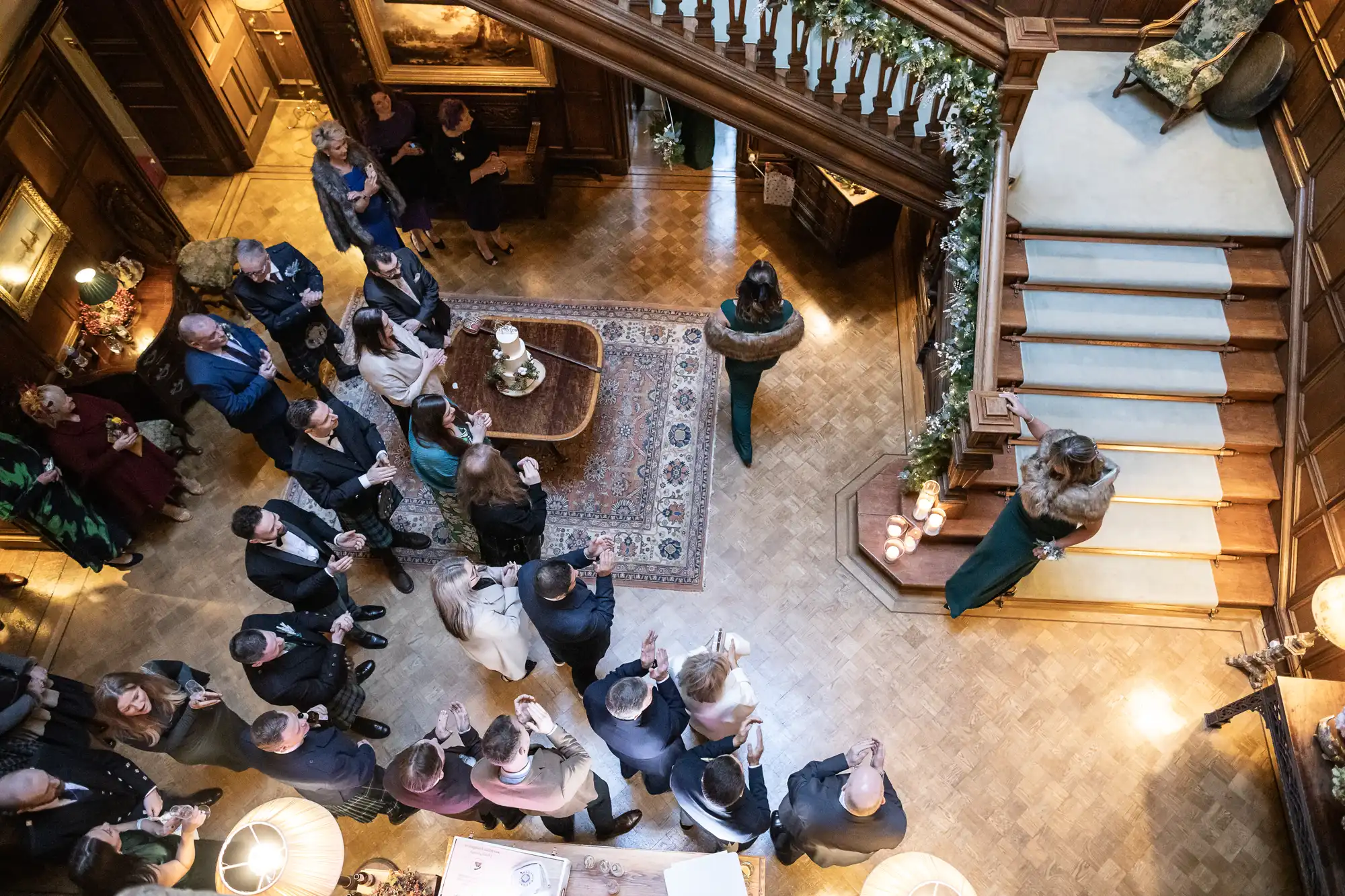 An overhead view of people gathered in a wood-paneled room near a staircase, some conversing, others looking at their phones. A decorated railing and a rug adorn the space.