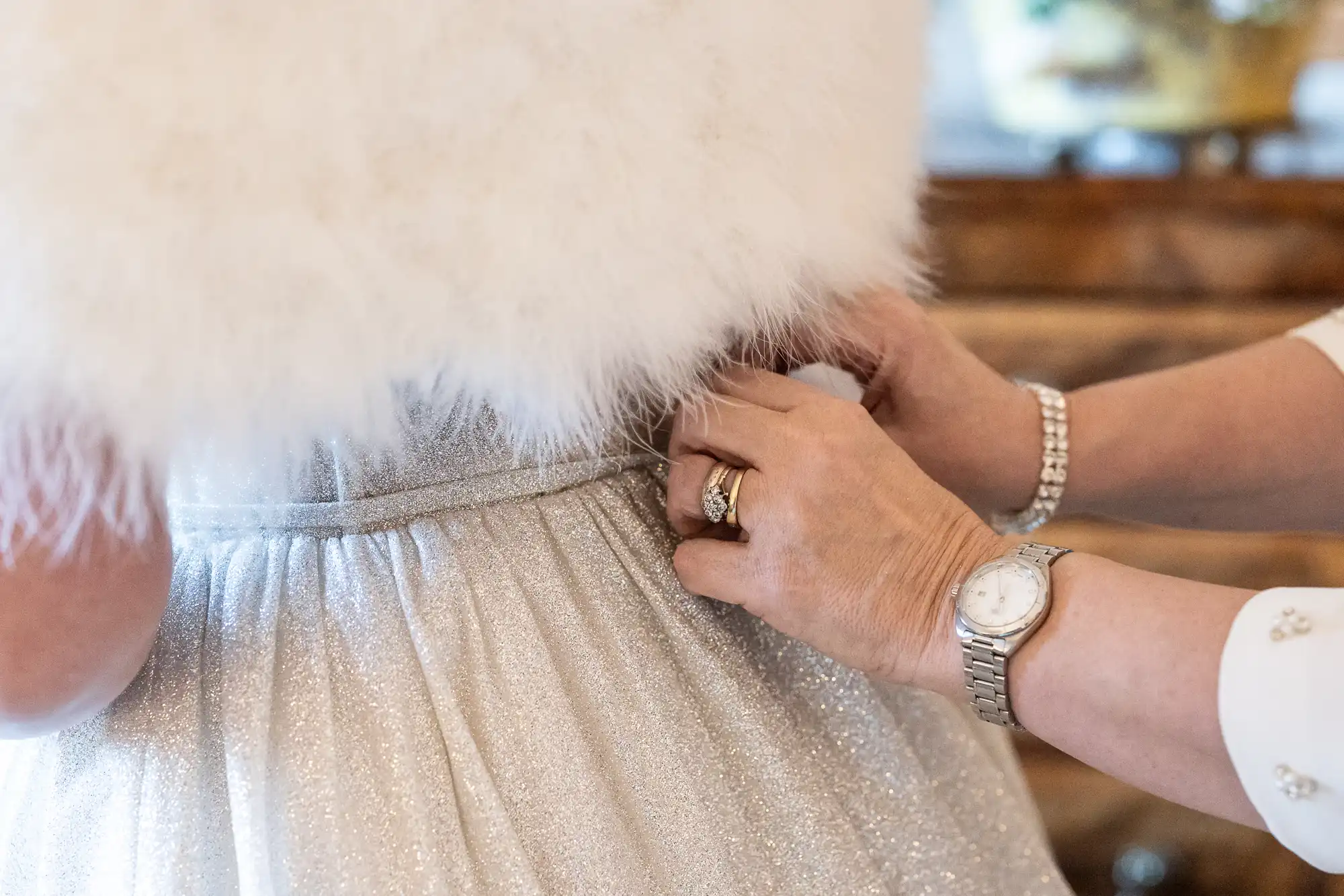 Close-up of two hands fastening the waist of a sparkly, silver dress with a fluffy white top. One hand has a ring and a watch, and the scene appears indoors.