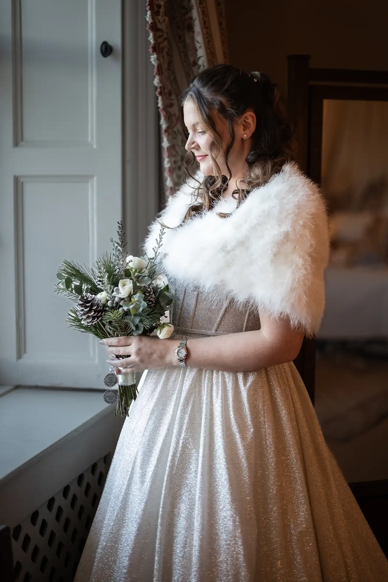 A woman in a white dress and fur shawl holds a bouquet of flowers while standing by a window.