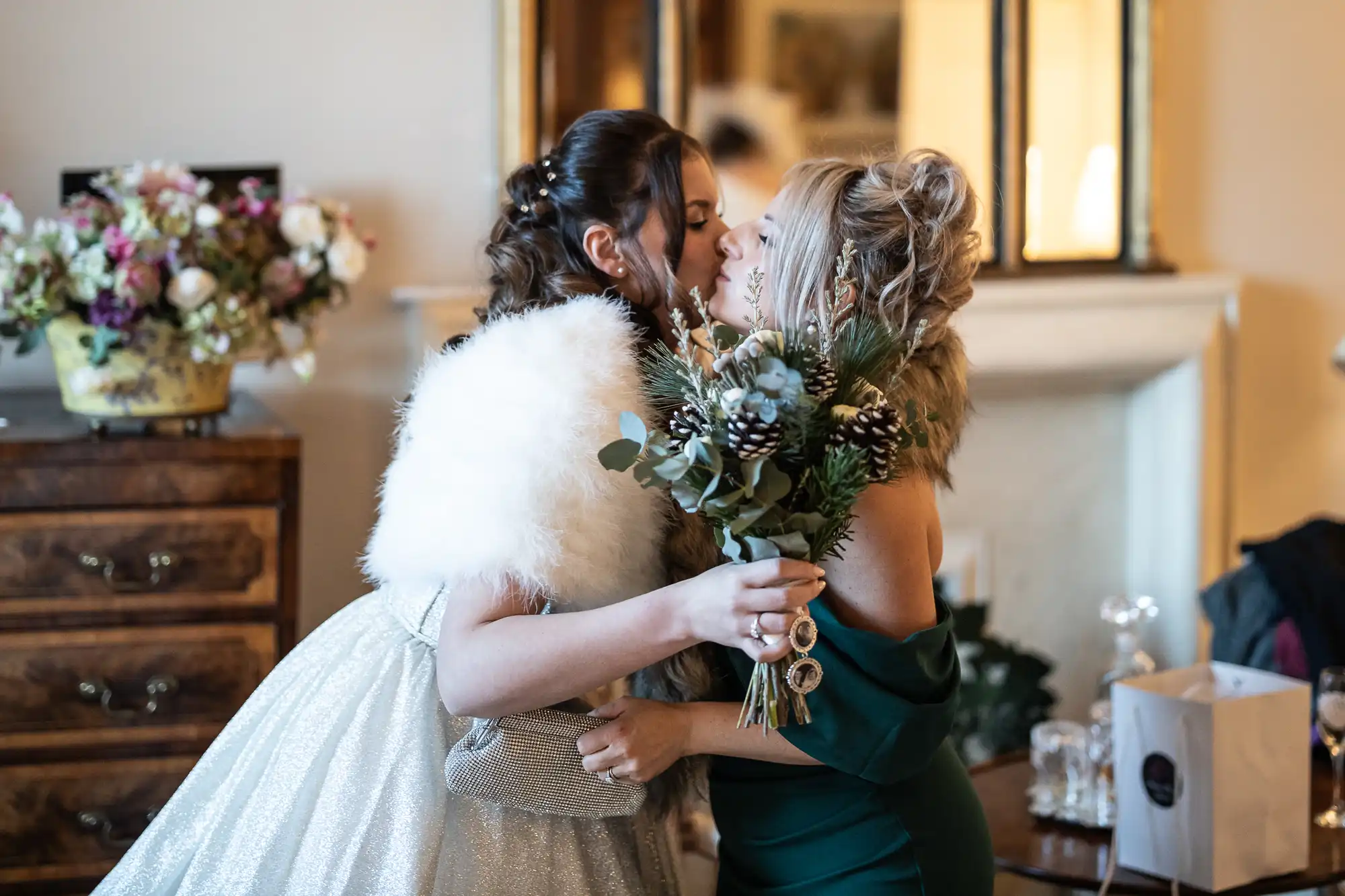 Two women dressed in formal attire share a kiss indoors near a decorative dresser with flowers. One holds a bouquet and the other is dressed in a fur shawl.