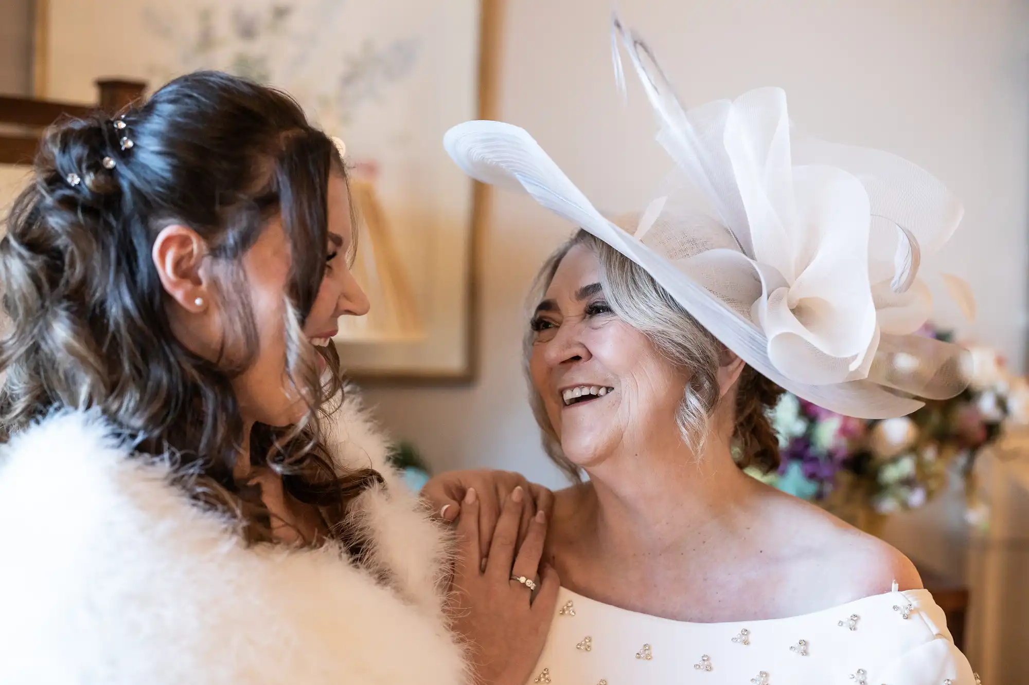 Two women smiling at each other. The younger woman wears a white furry wrap, and the older woman wears a white outfit with a large white hat. Background includes blurred floral decor.