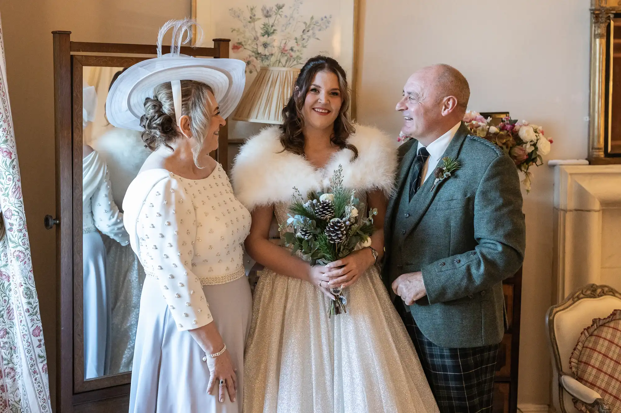 A bride in a white gown with a fur shawl stands between an older woman in a white dress and hat and an older man in a green suit, all smiling indoors next to a mirror and floral decor.