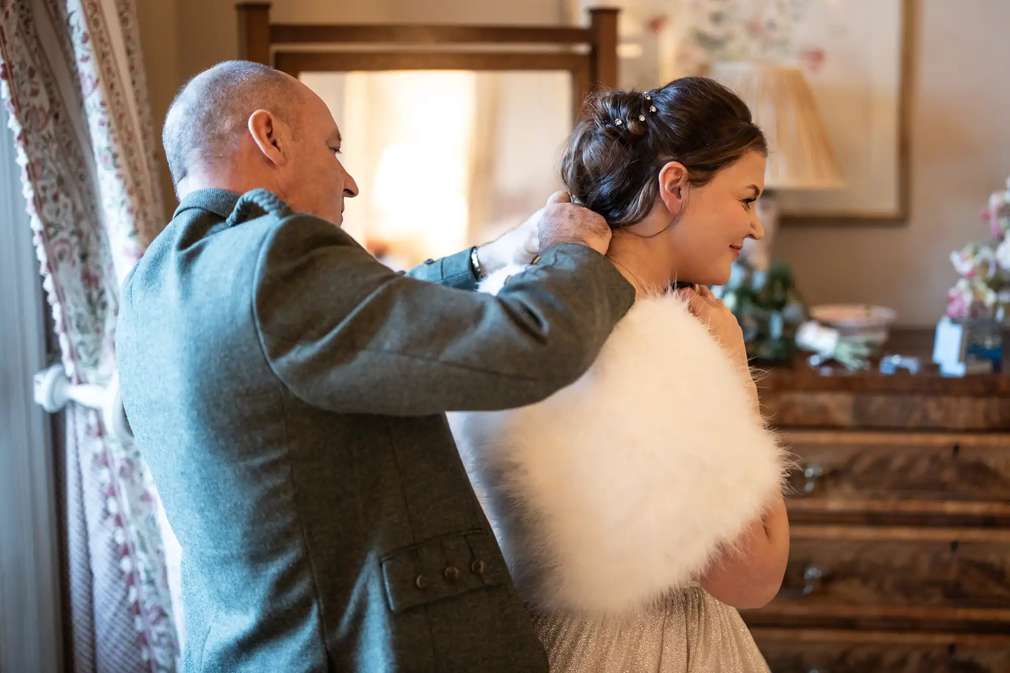 A man helps a woman with a white fur shawl, standing in a warmly lit room with a mirror and wooden furniture in the background.