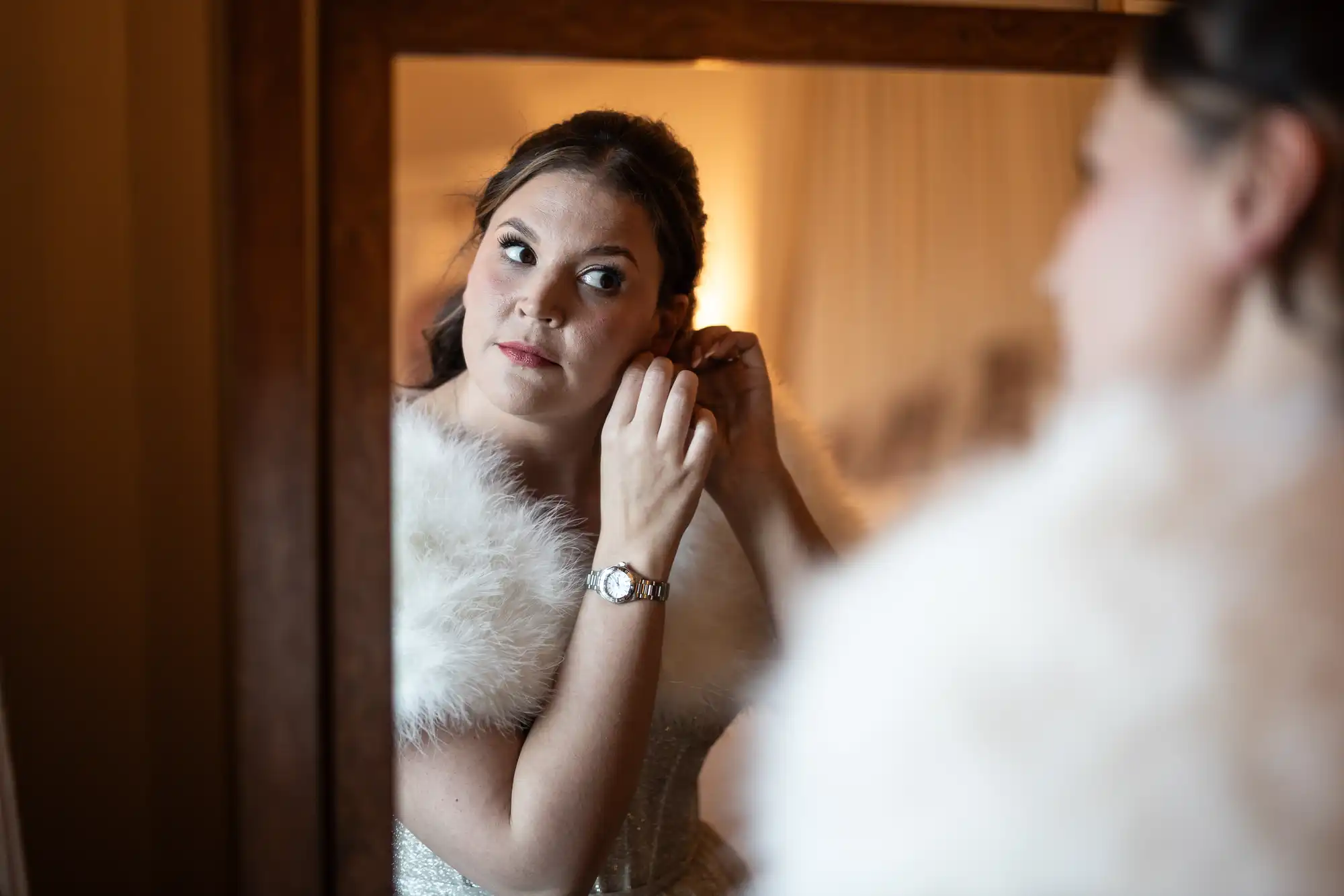 A woman is seen in a mirror putting on an earring, wearing a watch and a fur stole.