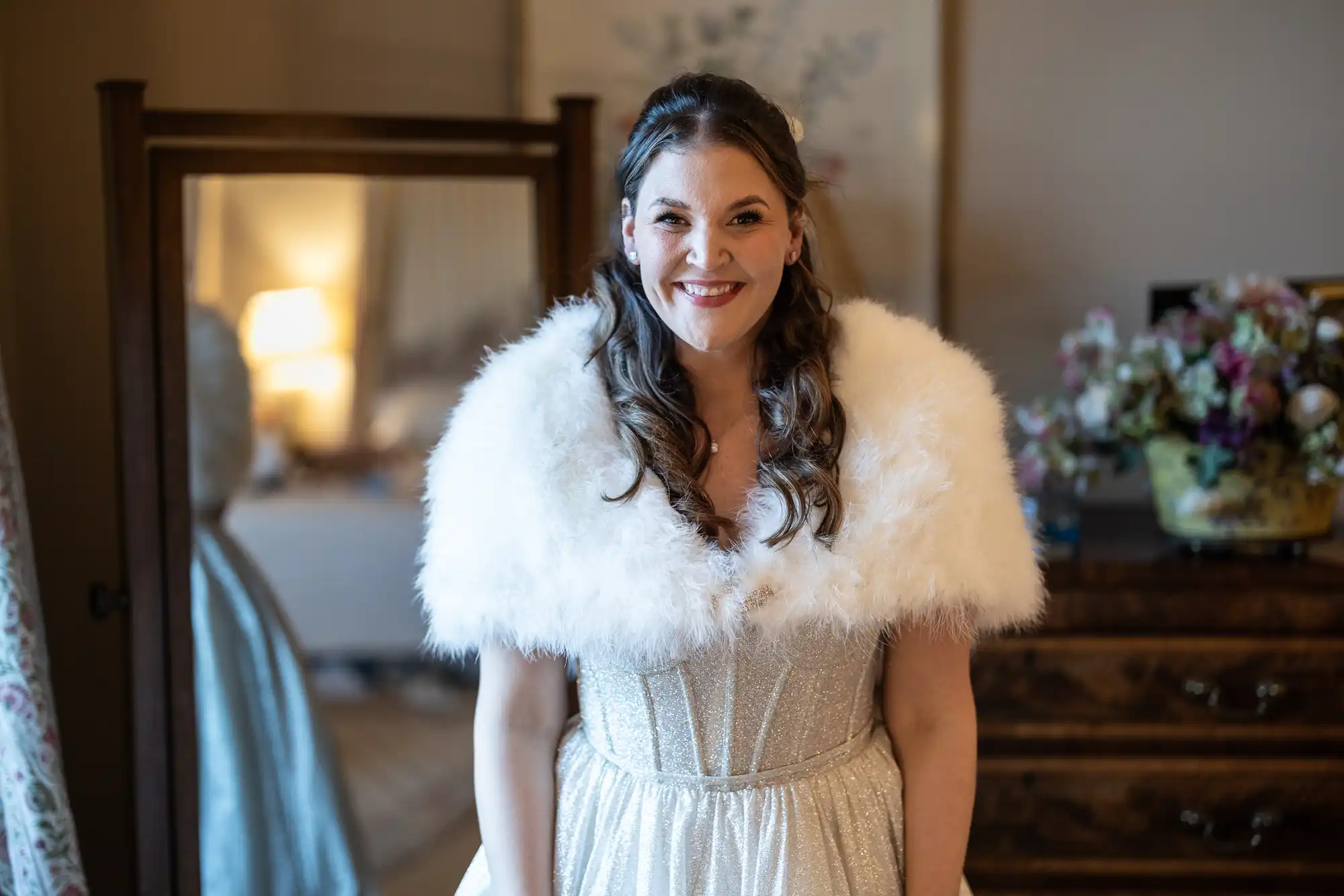 A woman in a sparkly dress and white fur stole smiles, standing in front of a dresser and mirror in a warmly lit room.