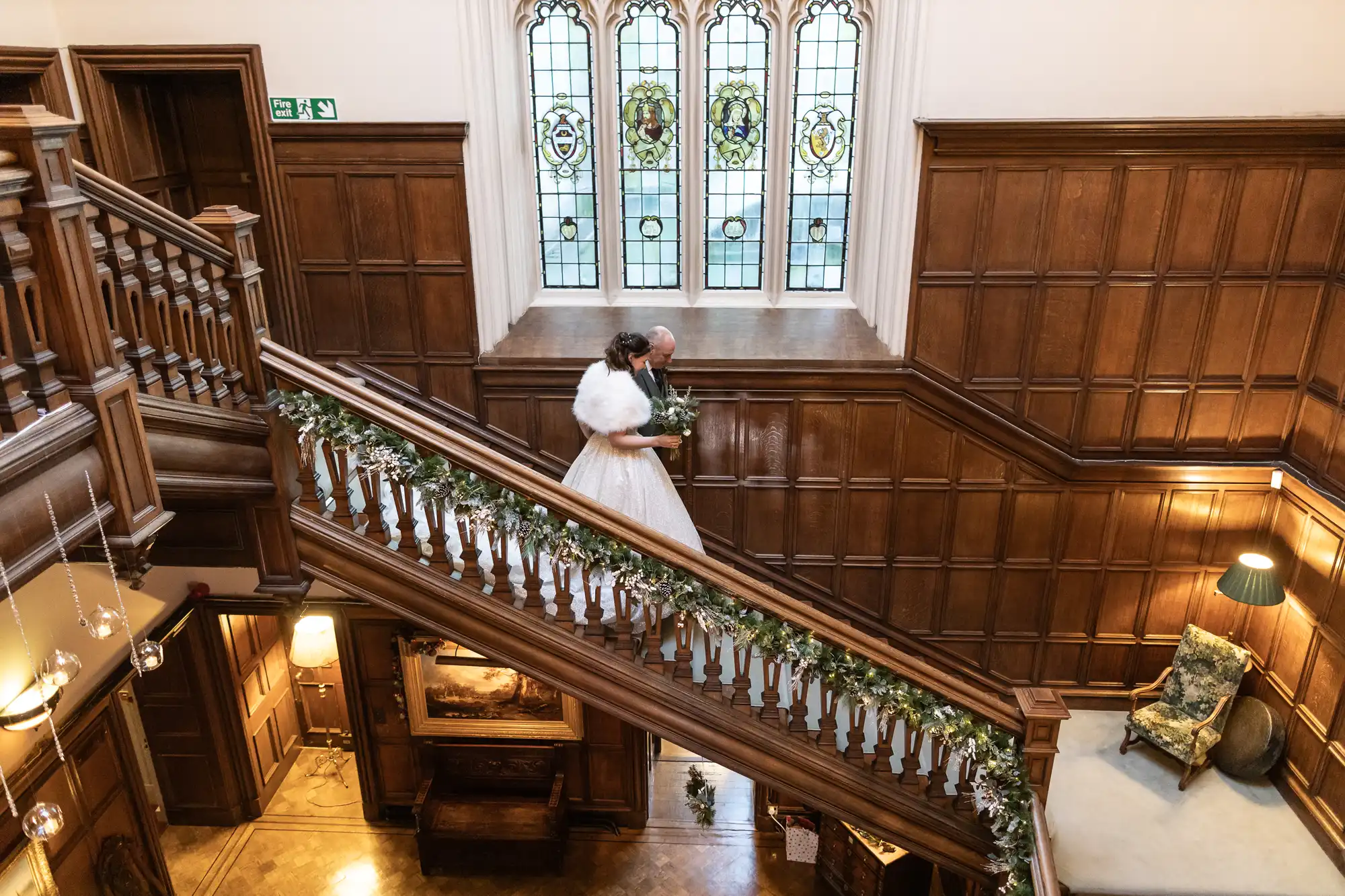 A bride and groom descend a wooden staircase adorned with greenery in a large, ornately decorated hall with stained glass windows.