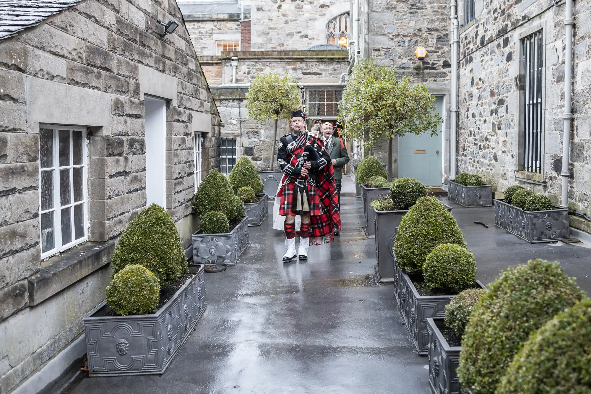 Two men wearing traditional Scottish kilts stand in a narrow courtyard with stone buildings and neatly trimmed bushes. A bagpiper leads the way, playing the bagpipes.