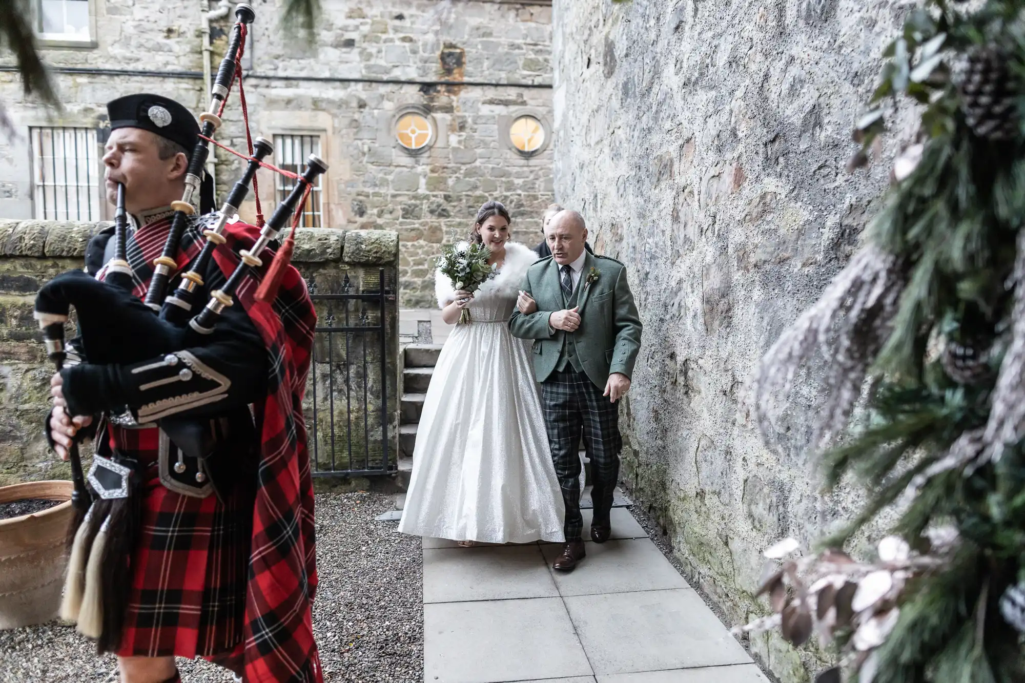 A bride holding a bouquet is escorted by an elderly man in a green jacket. They walk along a stone pathway with a bagpiper playing in the foreground, beside a stone wall and building.