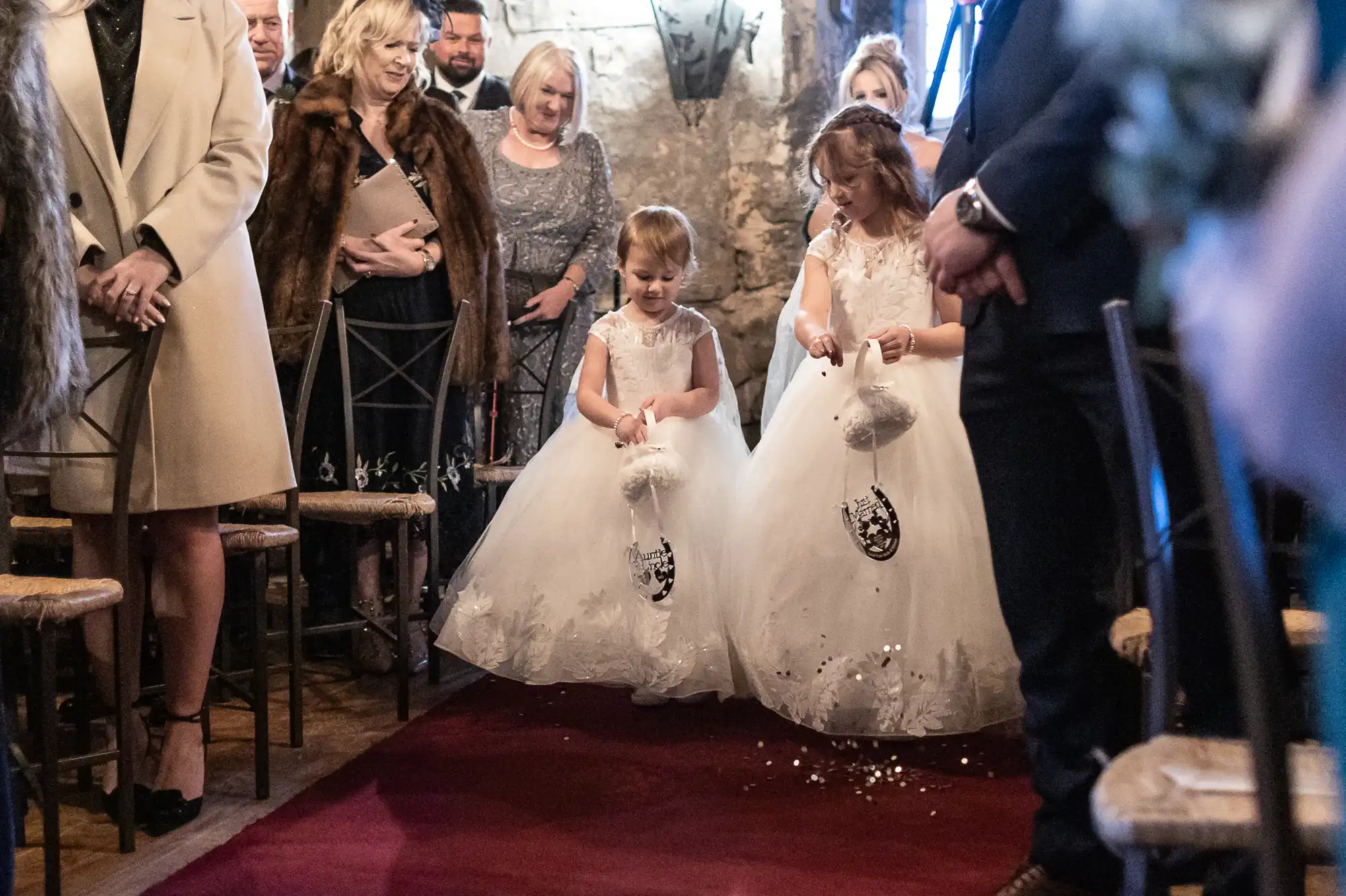 Two young girls in white dresses walk down an aisle, scattering flower petals, with seated guests watching them during a wedding ceremony.