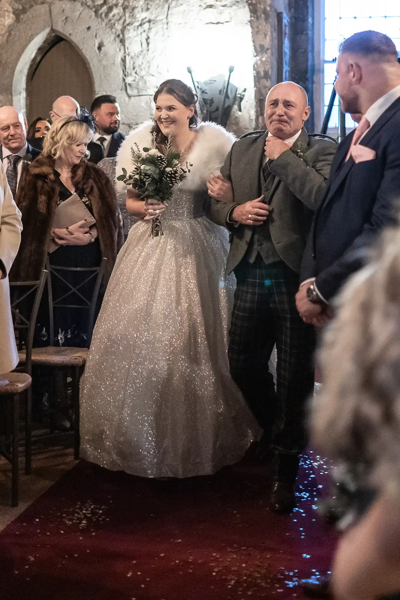 A bride in a sparkling gown with a fur shawl walks down the aisle with an elderly man holding her arm. Guests in formal attire watch from their seats.