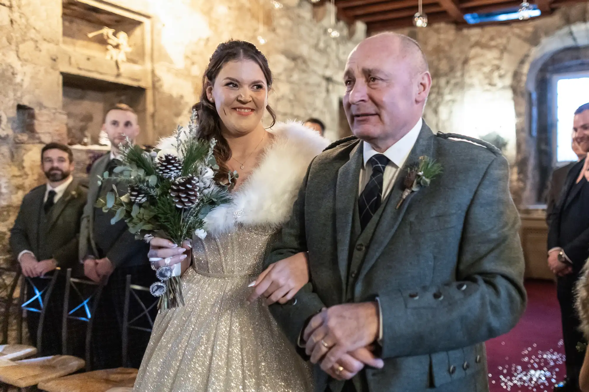 A bride in a sparkling dress and fur stole walks down the aisle with an older man in a gray suit, holding a flower bouquet, in a stone interior setting. Guests look on from both sides.