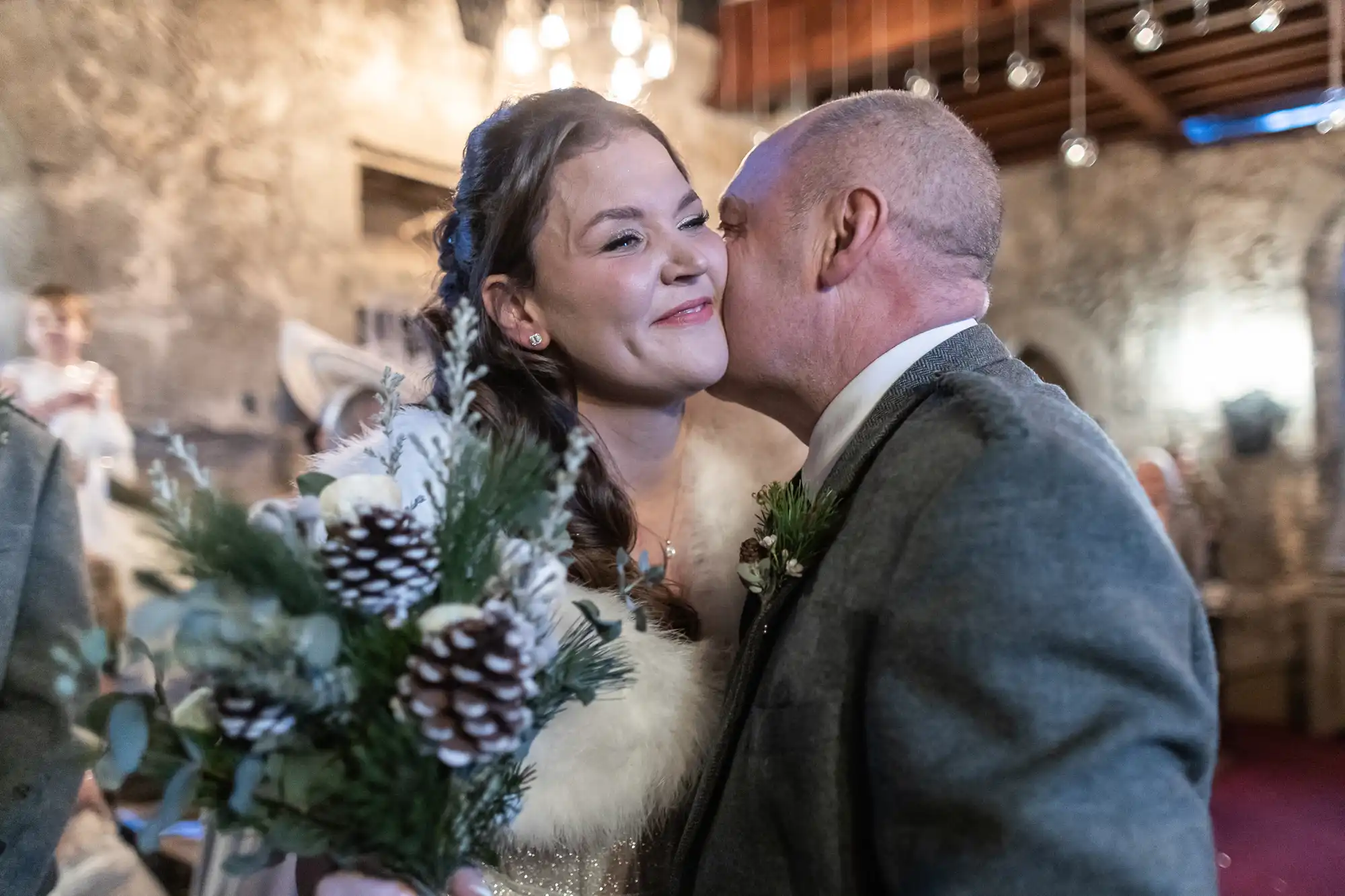 A man kisses a smiling woman on the cheek in a warmly lit room while she holds a bouquet adorned with pinecones and greenery.