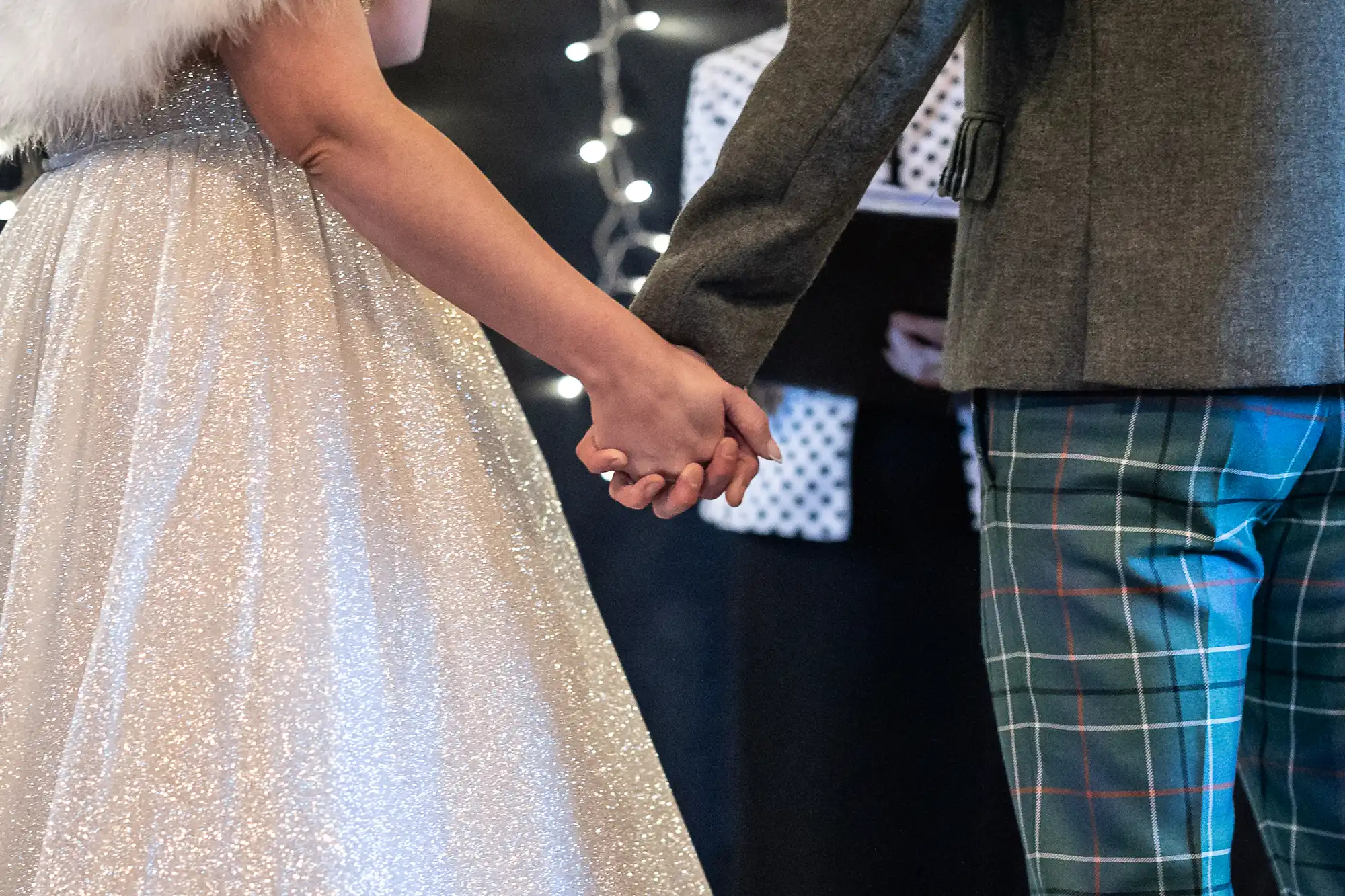 A couple holding hands, with the woman in a sparkling white dress and the man in a grey jacket and plaid pants.