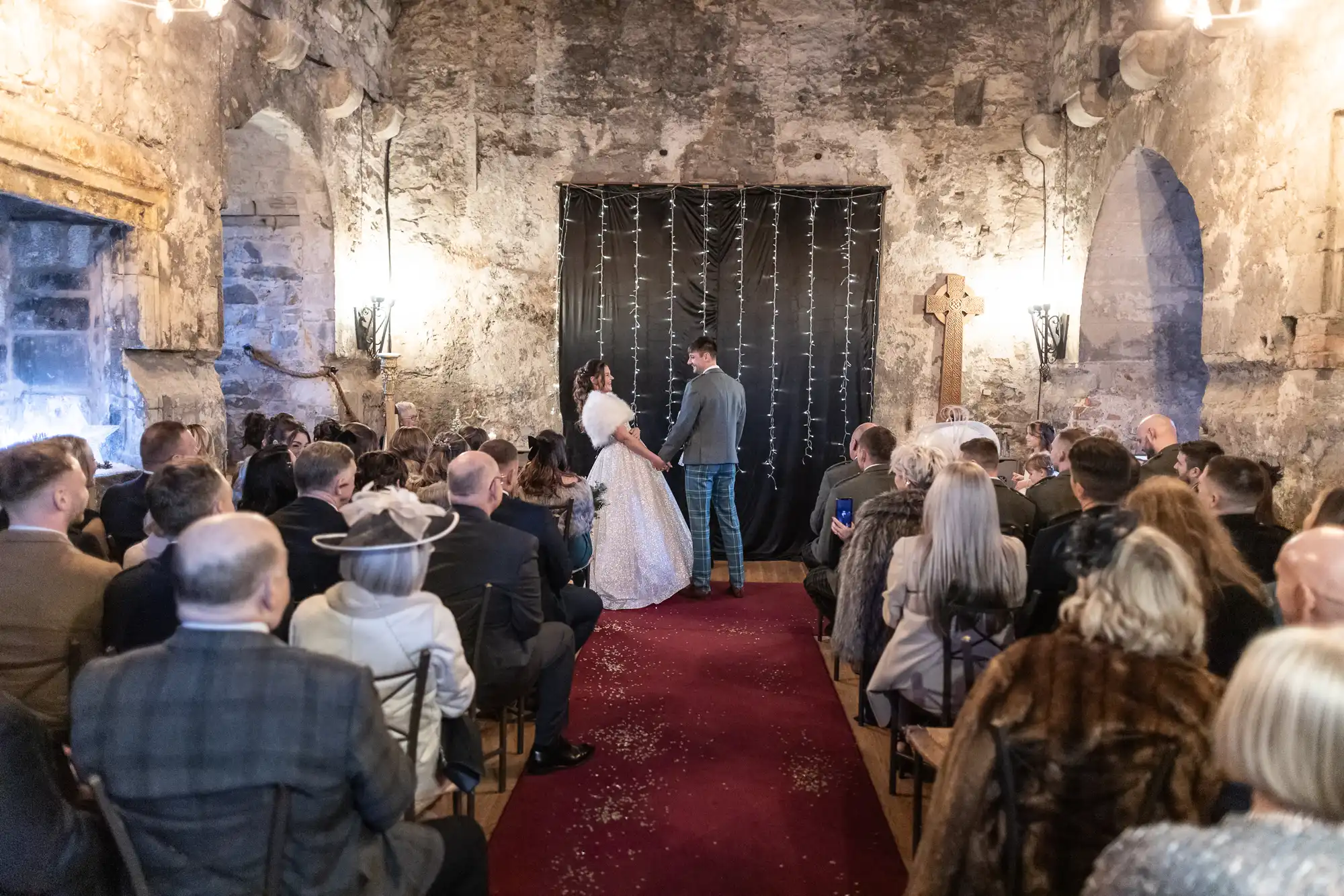 A couple stands facing each other at the altar in front of a congregation during a wedding ceremony in a rustic stone-walled setting.