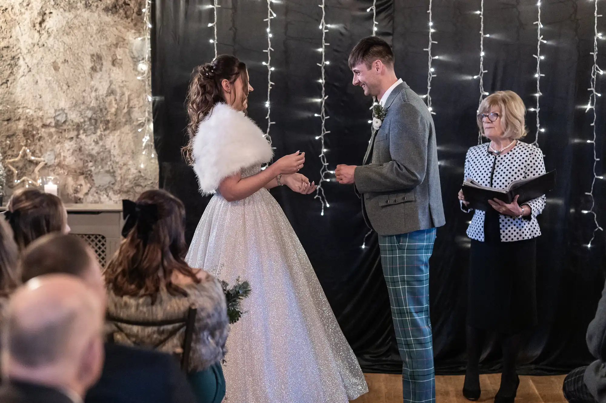 A couple exchanges rings during a wedding ceremony. The bride wears a white gown and fur shawl, while the groom wears a grey jacket and plaid trousers. They are joined by an officiant and guests are seated nearby.