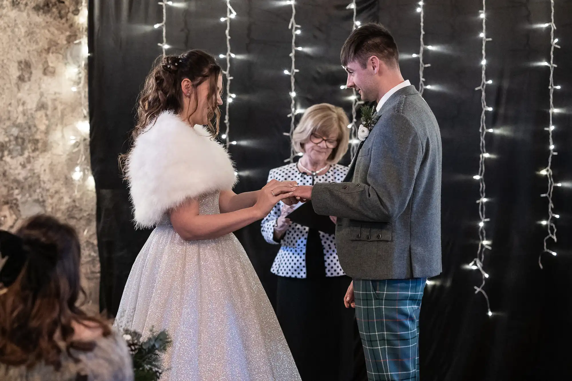 A couple exchanges rings in front of an officiant during their wedding ceremony. String lights hang in the background, adding a festive touch.