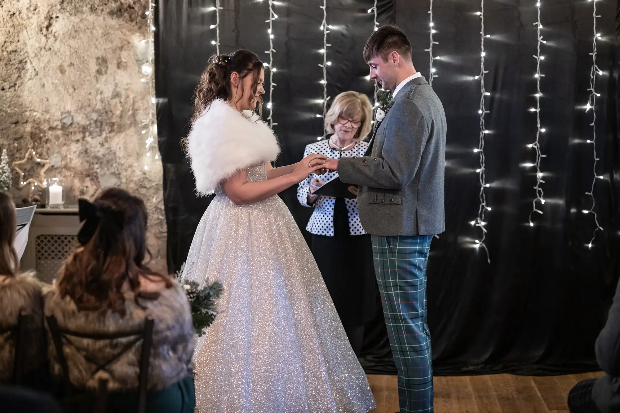 A bride and groom exchange rings during their wedding ceremony. The officiant stands behind them. They are in front of a dark curtain decorated with string lights.