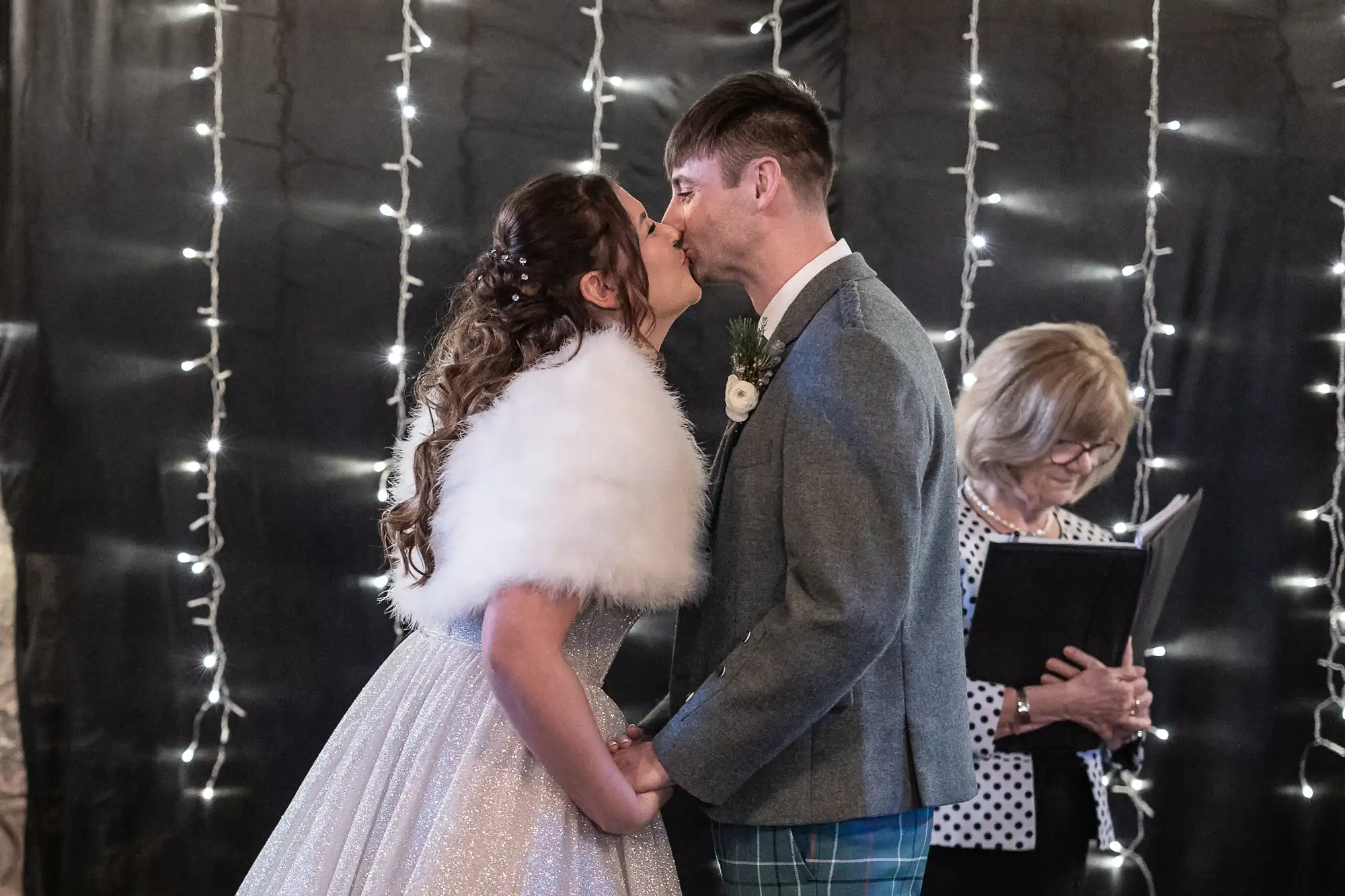 A bride and groom share a kiss while holding hands during their wedding ceremony with a woman reading from a book in the background.