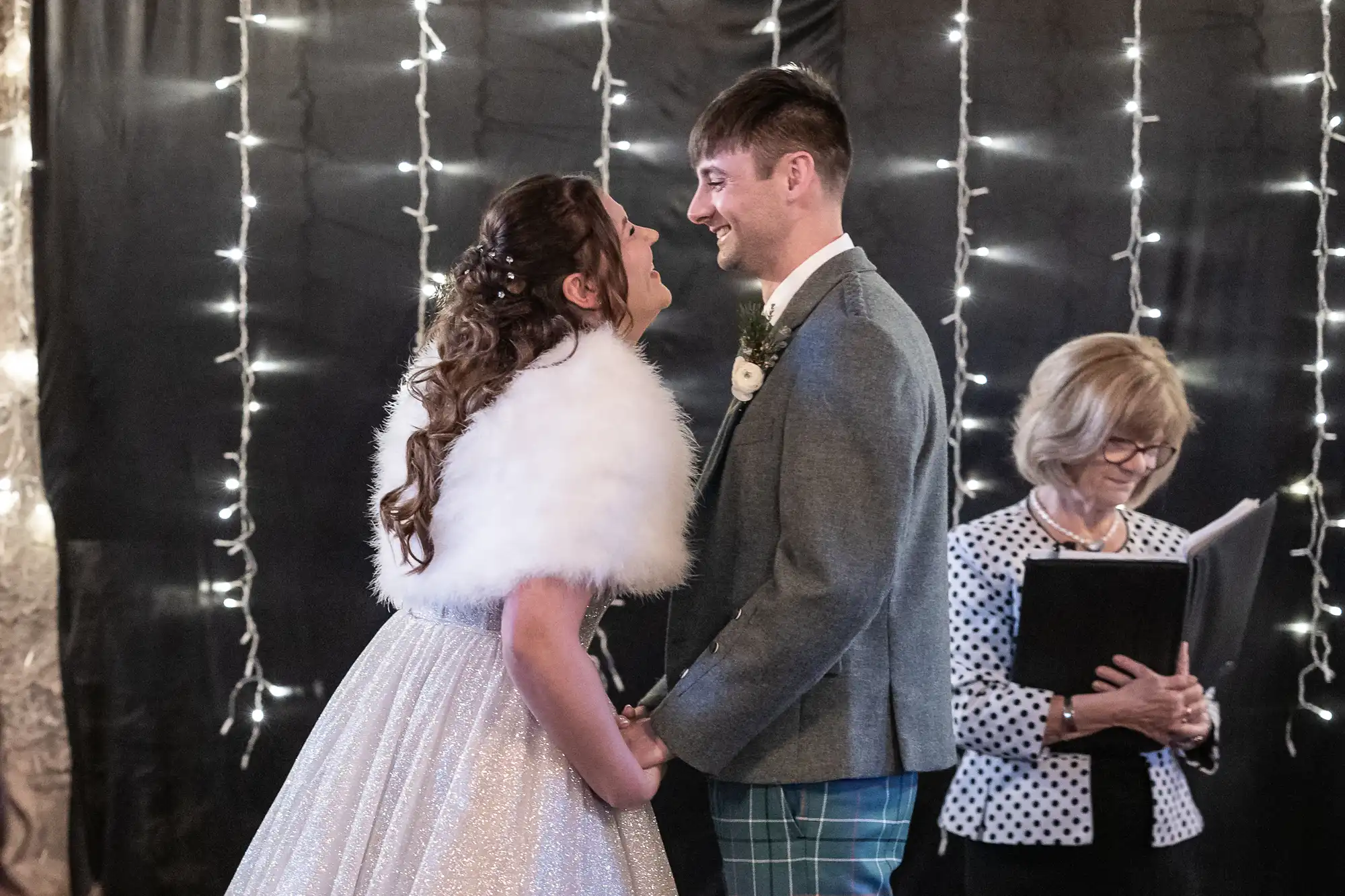 A couple stands facing each other and holding hands, smiling under string lights while an officiant with a book stands nearby.