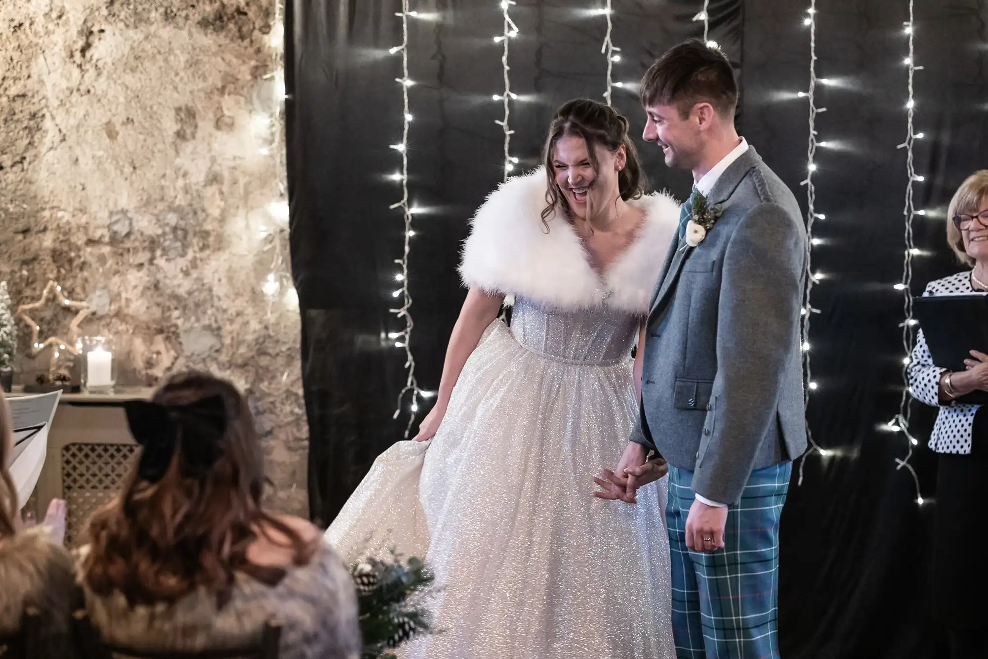 A bride and groom hold hands and smile during their wedding ceremony, with string lights and black drapery as a backdrop. Guests and an officiant are visible in the background.