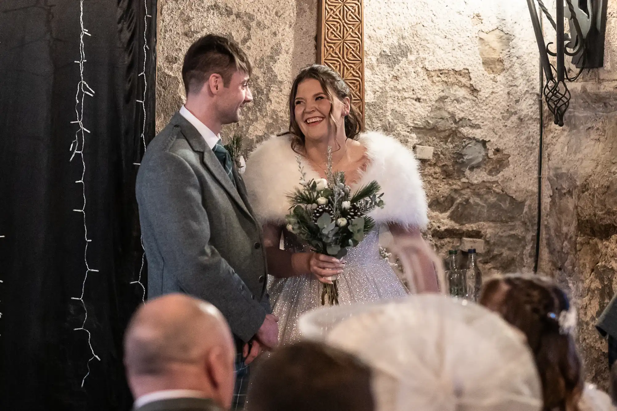 A man and woman stand together, smiling, at what appears to be a wedding ceremony. The woman is holding a bouquet and is dressed in a white gown with a fur shawl.