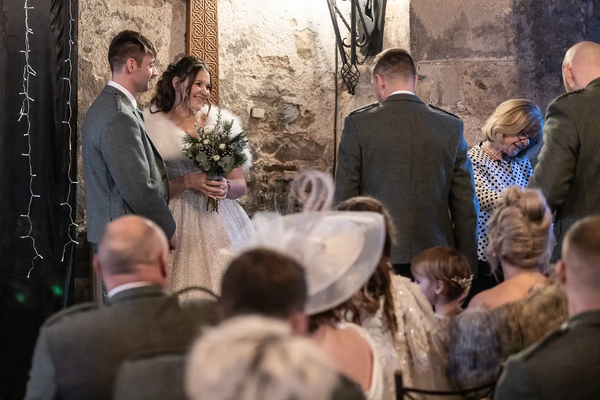 A bride and groom stand smiling during a wedding ceremony in a rustic venue surrounded by family and guests. A woman in a polka-dot top holds a book beside them.