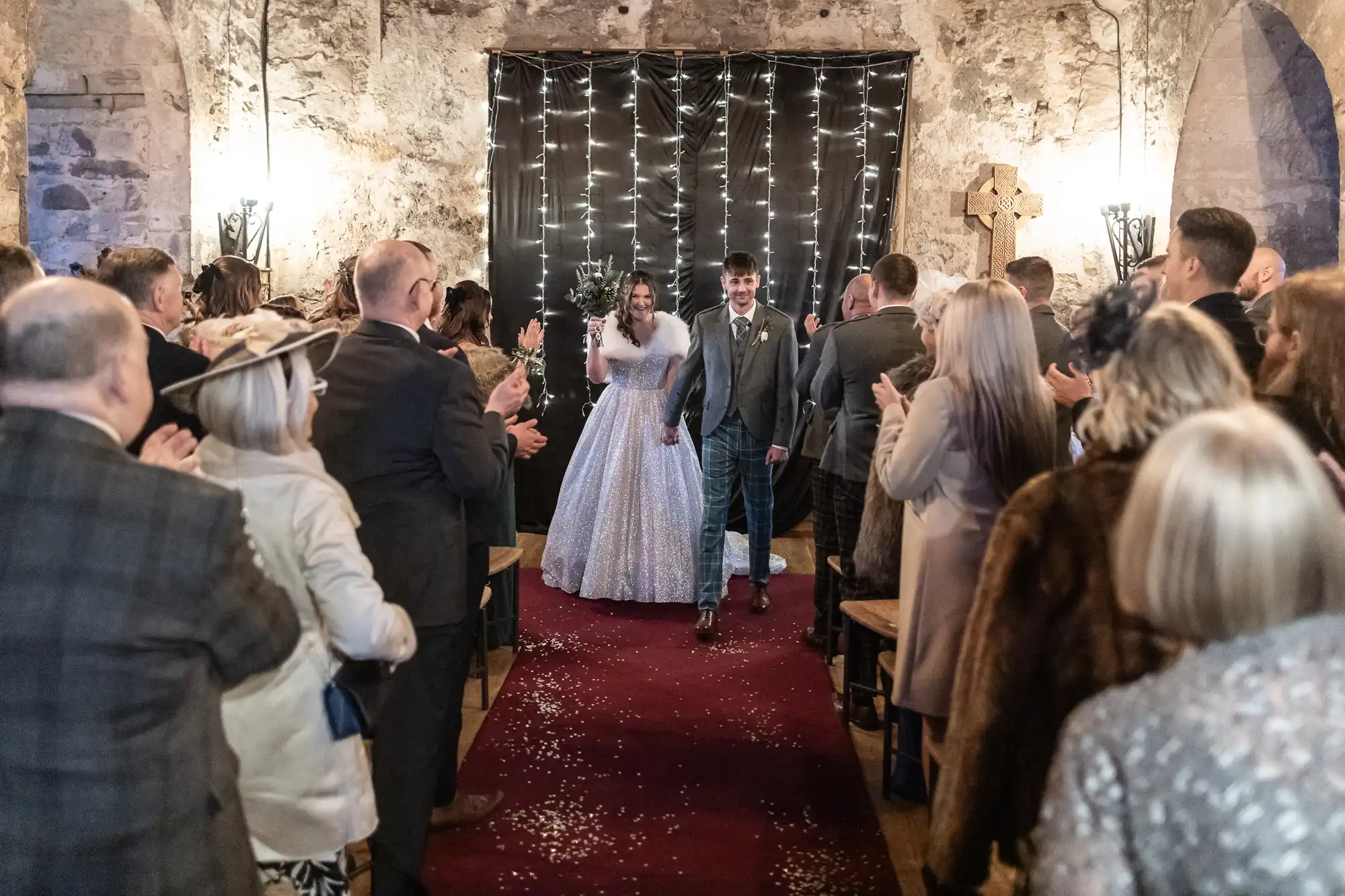 A newlywed couple walks down a red carpeted aisle in a rustic venue, with guests clapping and celebrating on either side. The bride wears a white gown, and the groom is in a suit. Twinkling lights hang behind them.