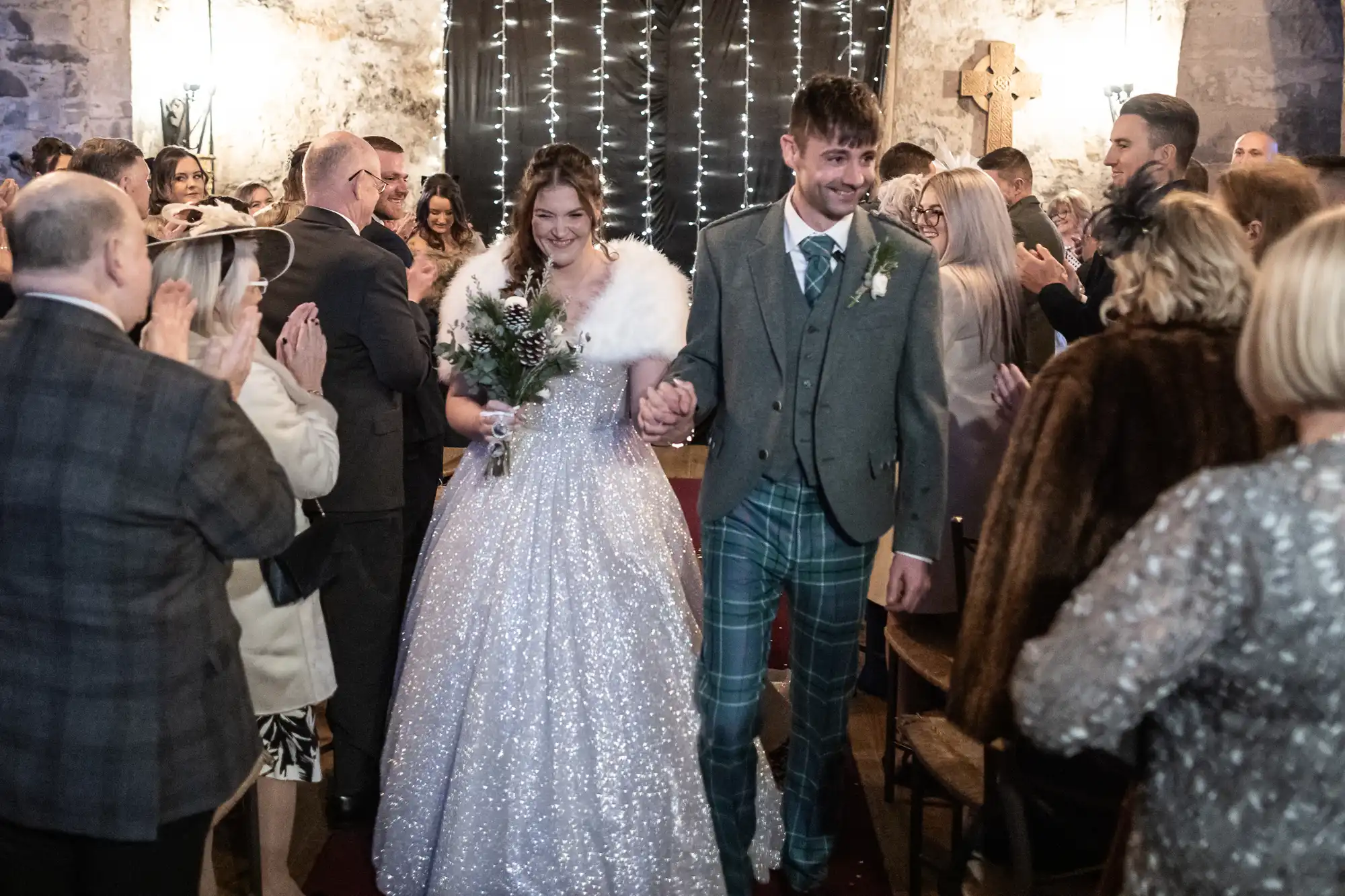 A bride and groom walk down the aisle surrounded by applauding guests. The bride wears a sparkling gown with a fur wrap and holds a bouquet. The groom wears a green suit with plaid trousers.