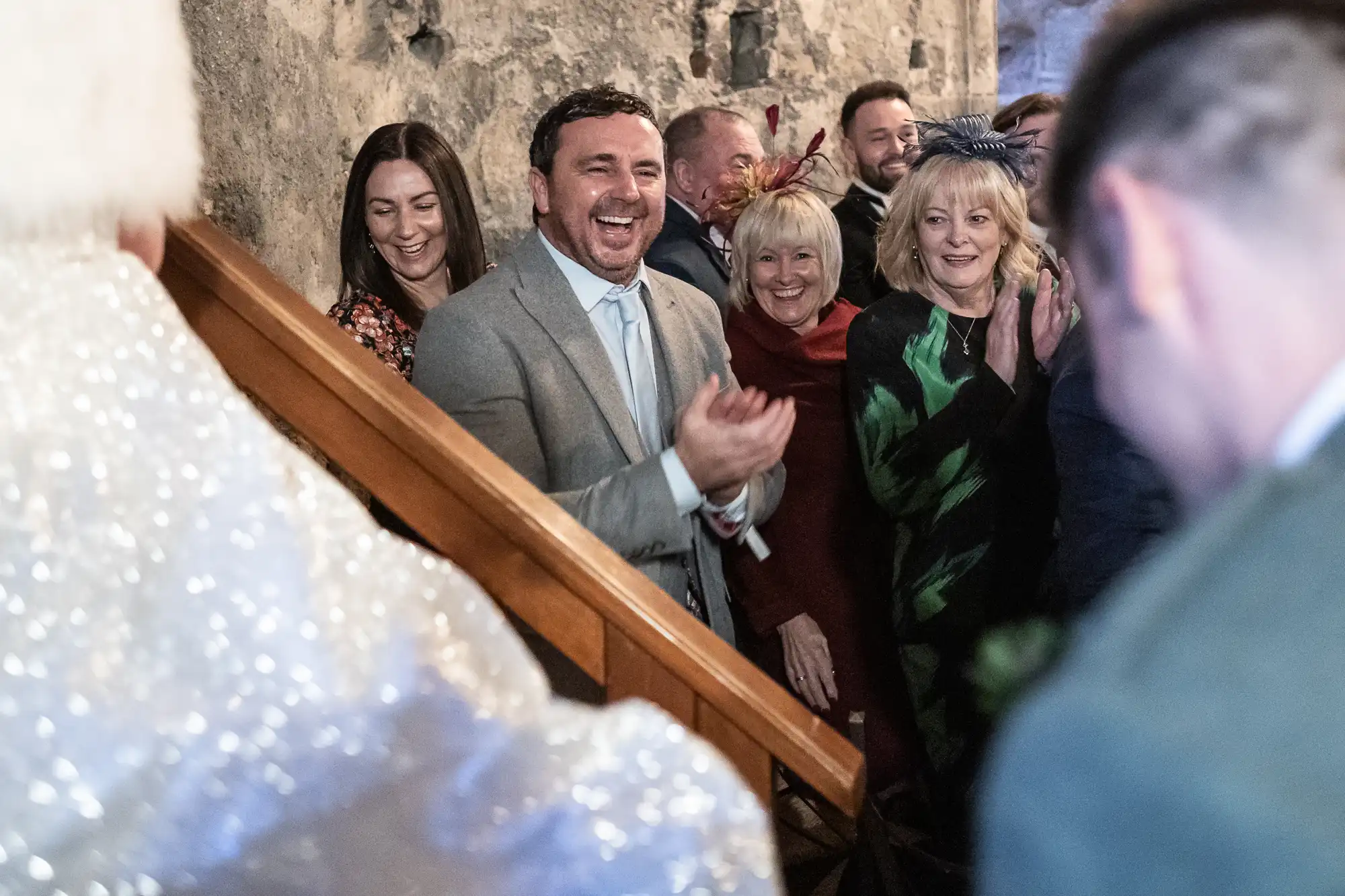 A group of people stands by a stone wall, clapping and smiling at an event. A man in a grey suit and tie is prominently seen in the center, looking towards the focus of their attention.