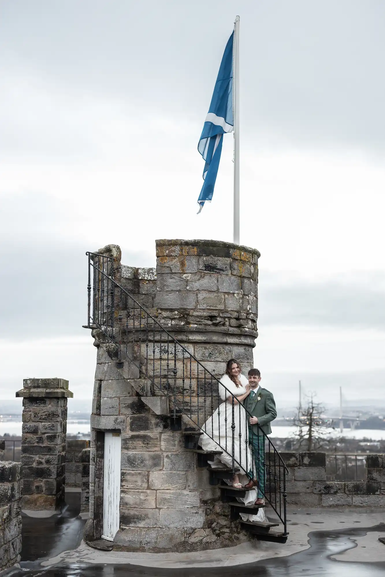 A couple stands on a spiral stone staircase of a historic structure with a blue flag flying at the top, surrounded by a cloudy sky and distant landscape.