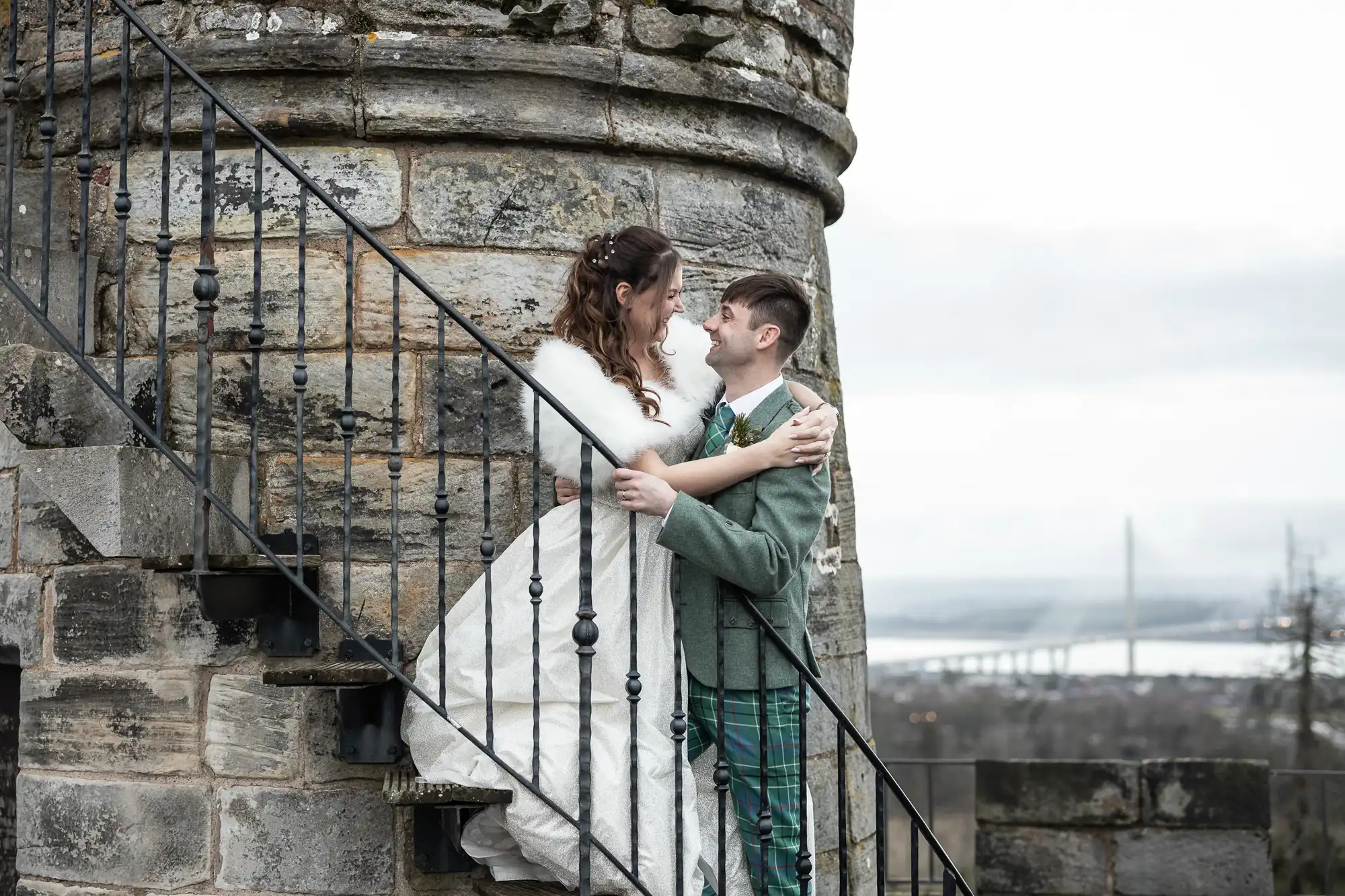 A bride in a white gown stands on an outdoor staircase, holding hands with a groom in green plaid pants and a grey jacket, both smiling in front of a stone tower and a distant bridge.