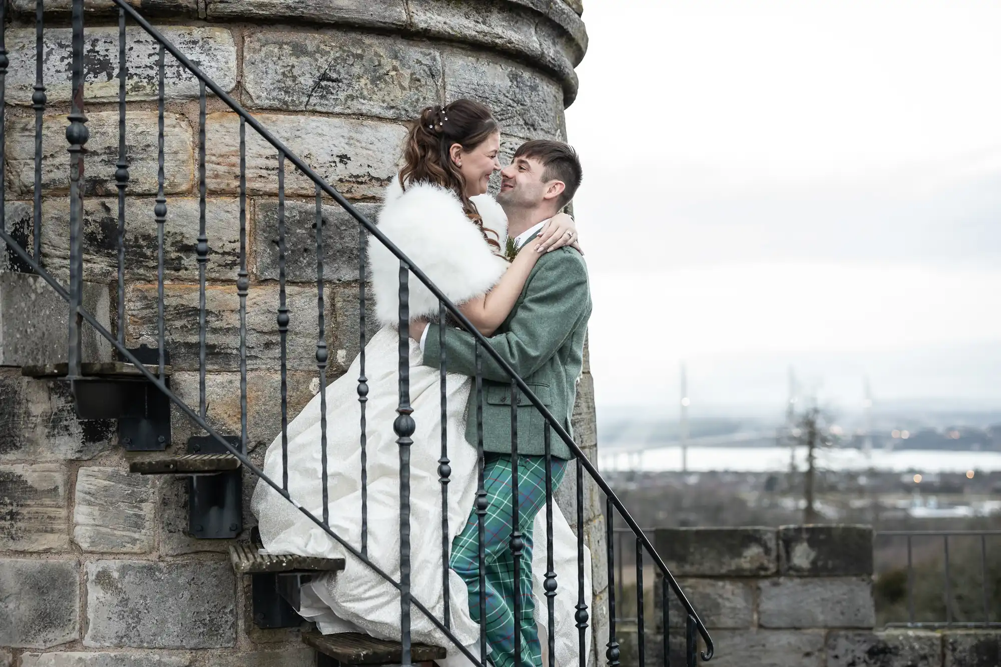 A couple embraces on an outdoor stone staircase, with the groom wearing a plaid suit and the bride in a white dress and fur shawl, while a distant landscape is visible in the background.