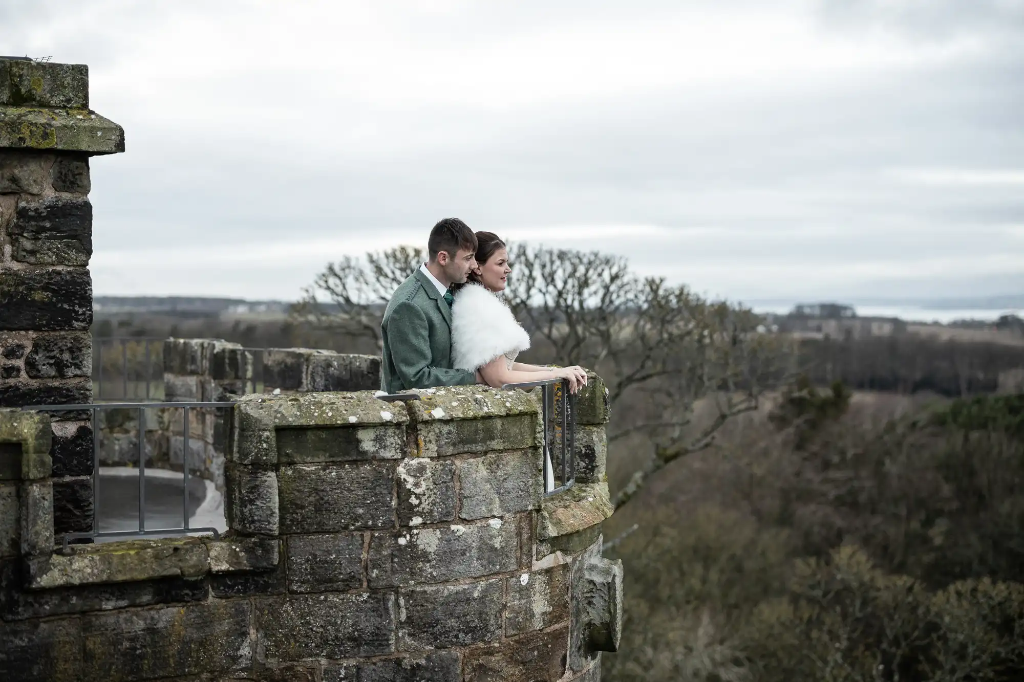 A couple stands together on the stone balcony of a castle, overlooking a vast landscape of trees and rolling hills under a cloudy sky.