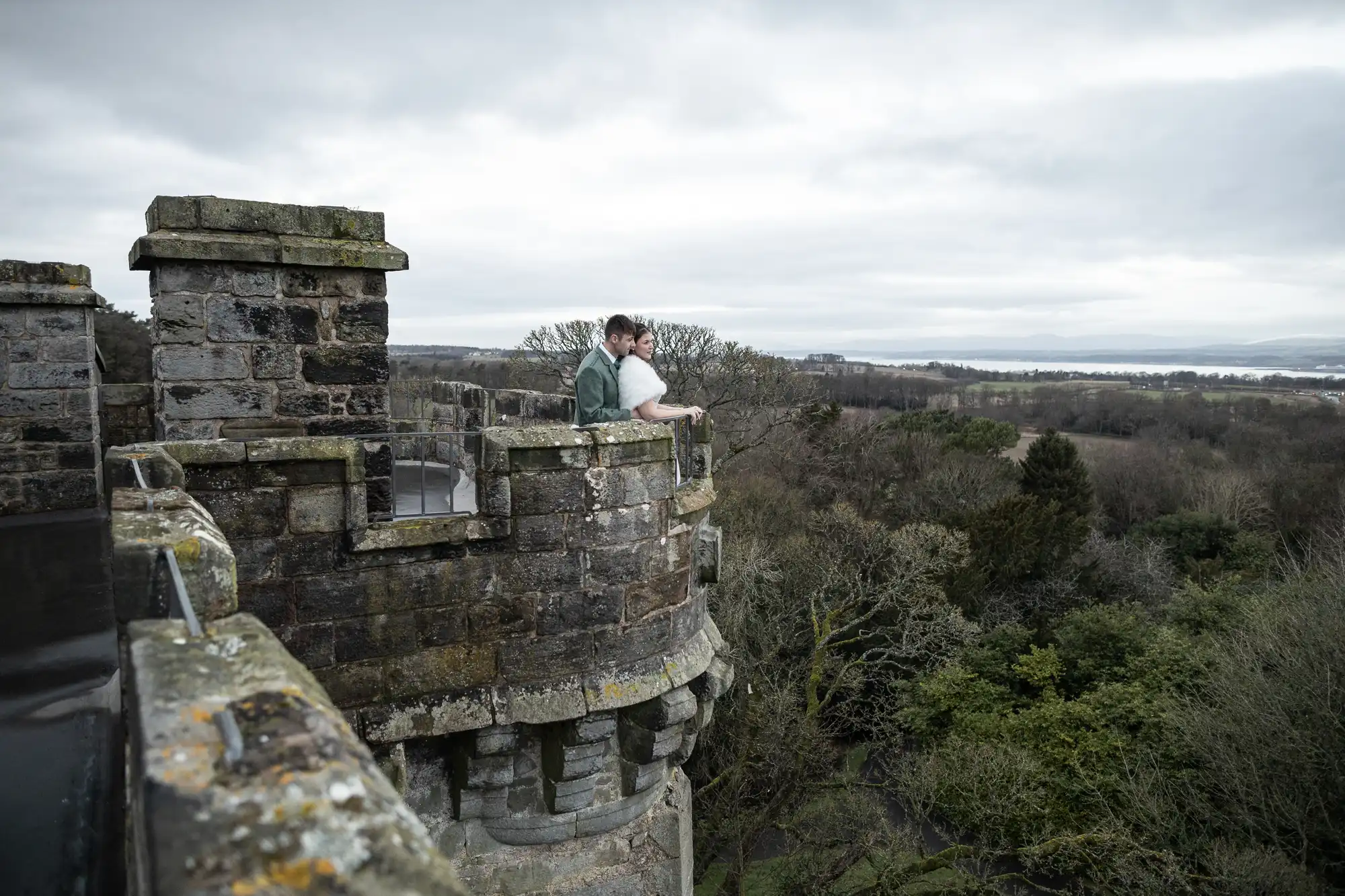 Two people stand on the lookout of a stone castle tower, gazing at the surrounding forest and distant landscape under a cloudy sky.