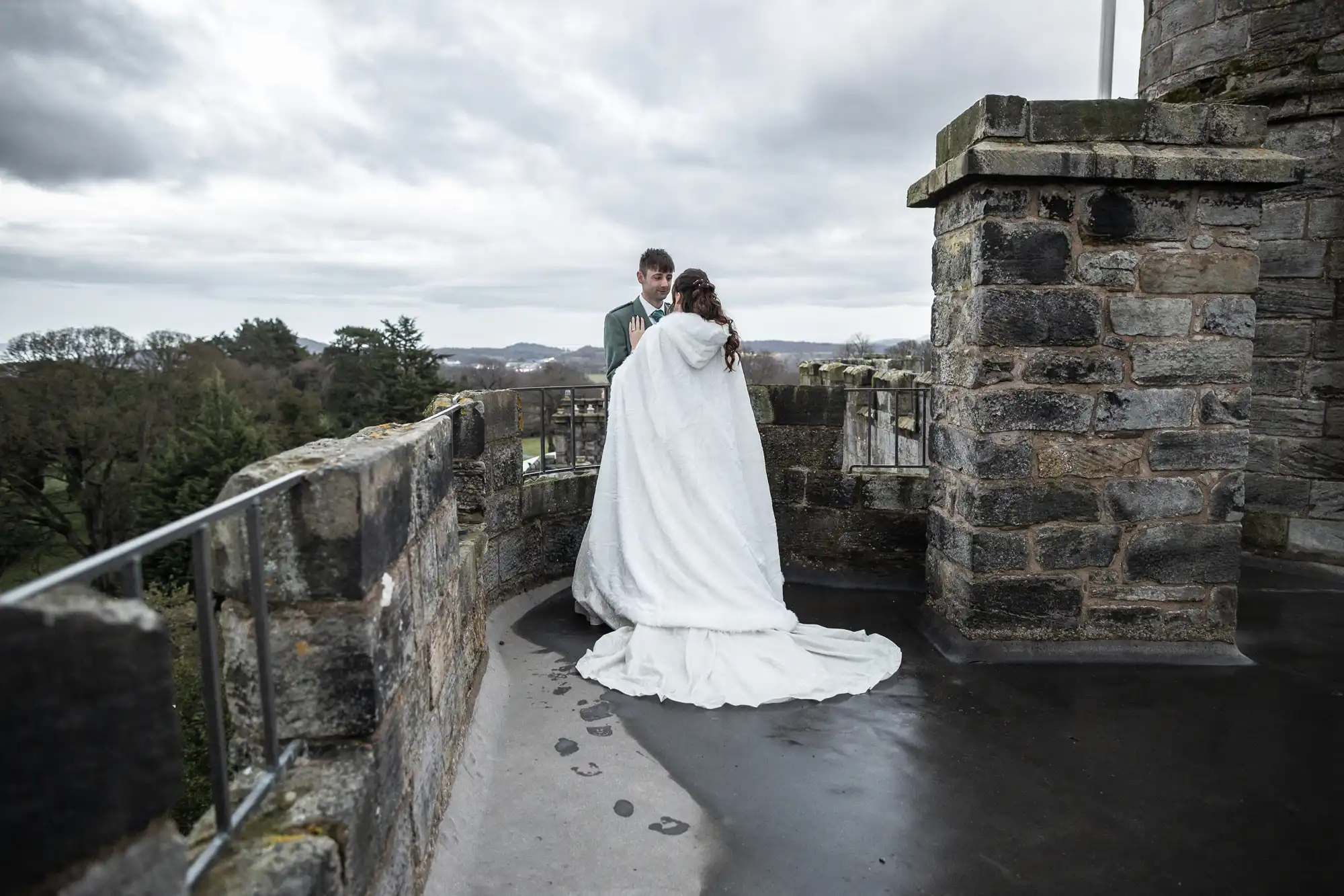 A bride and groom stand together on a stone balcony overlooking a cloudy landscape. The bride is wrapped in a white cloak, and there are footprints visible on the wet ground.