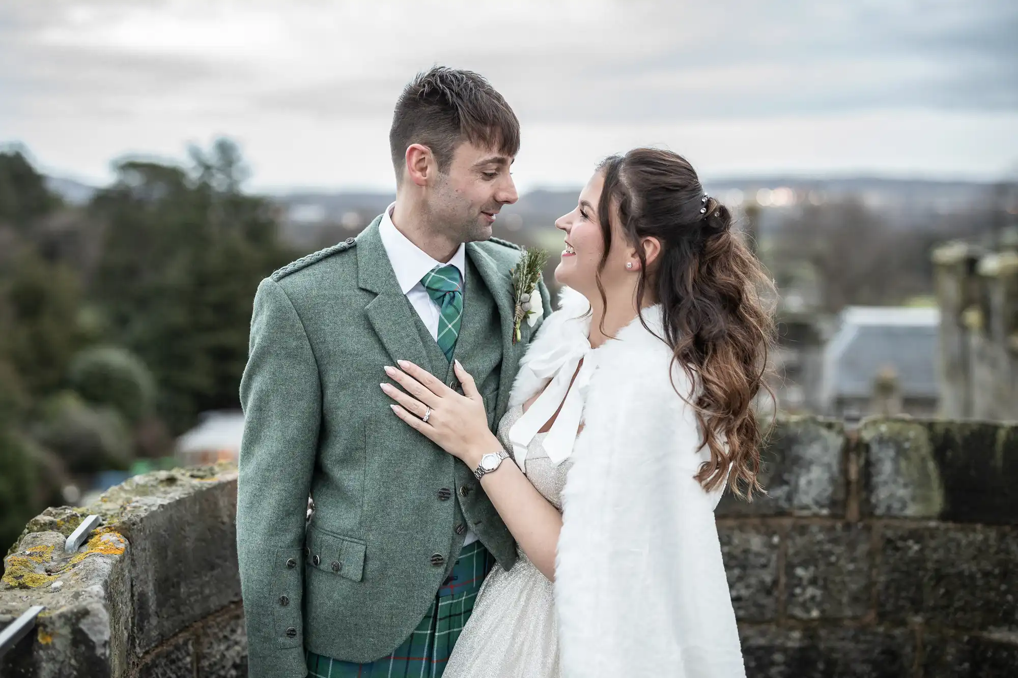 A couple dressed in formal wear gazes at each other while standing outdoors on a stone structure. The man wears a green suit with a tartan kilt; the woman is in a white dress with a fur shawl.