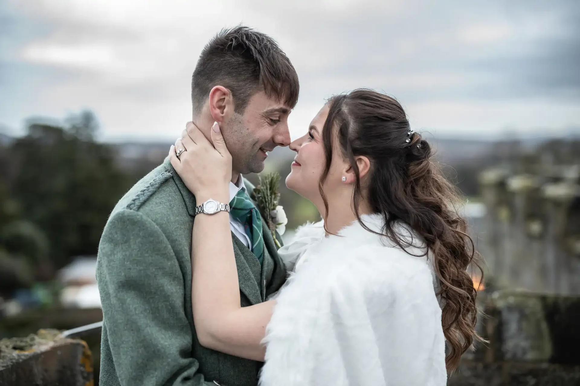 wedding photographer at Dundas Castle: a couple wearing wedding attire gazes into each other's eyes and smiles, standing outdoors with a blurred natural background. The man wears a gray suit and green tie; the woman wears a white fur shoulder wrap.